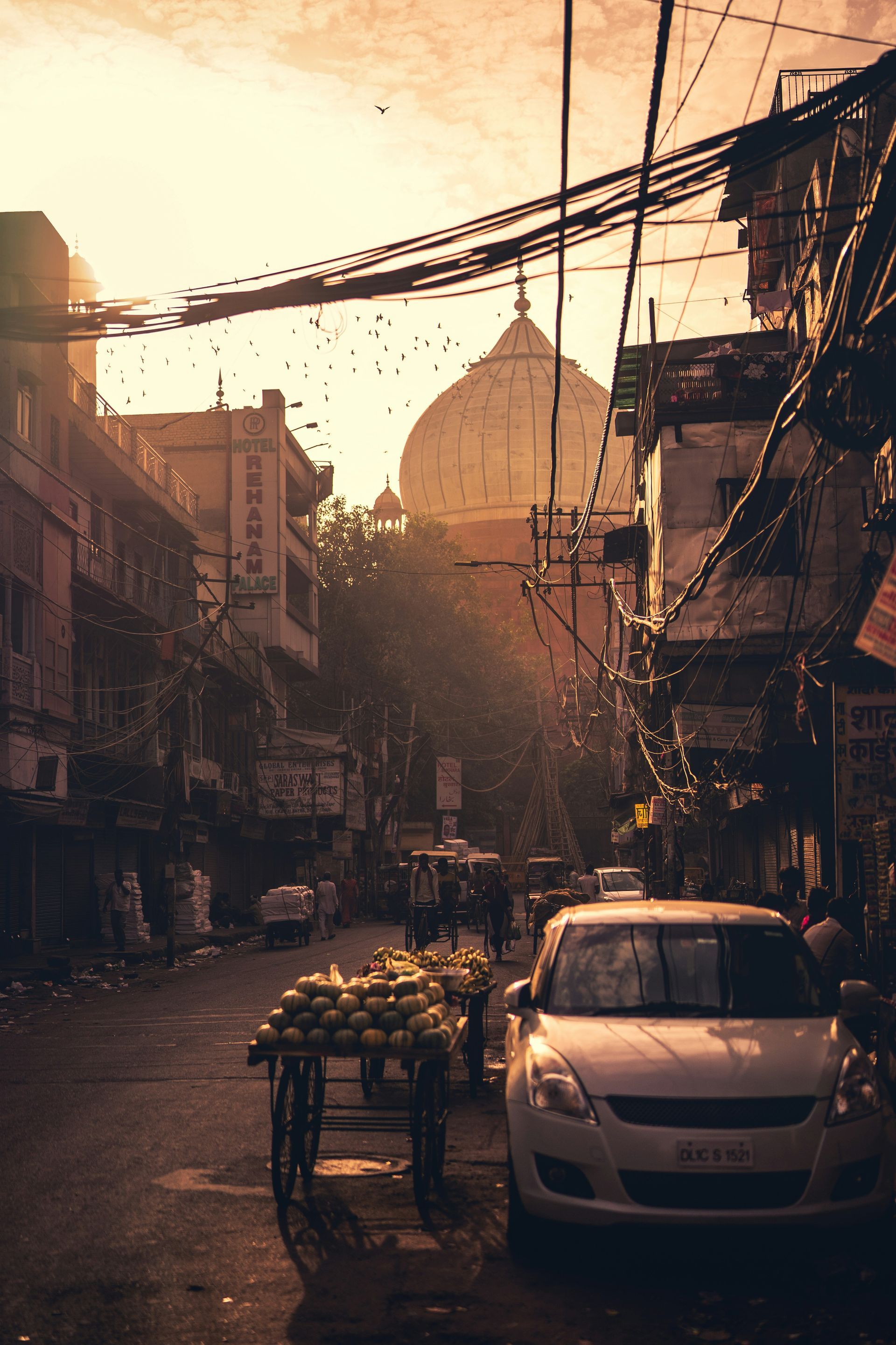 A sunlit street in New Delhi, India, with a domed building in the background; a car and fruit cart in the foreground.