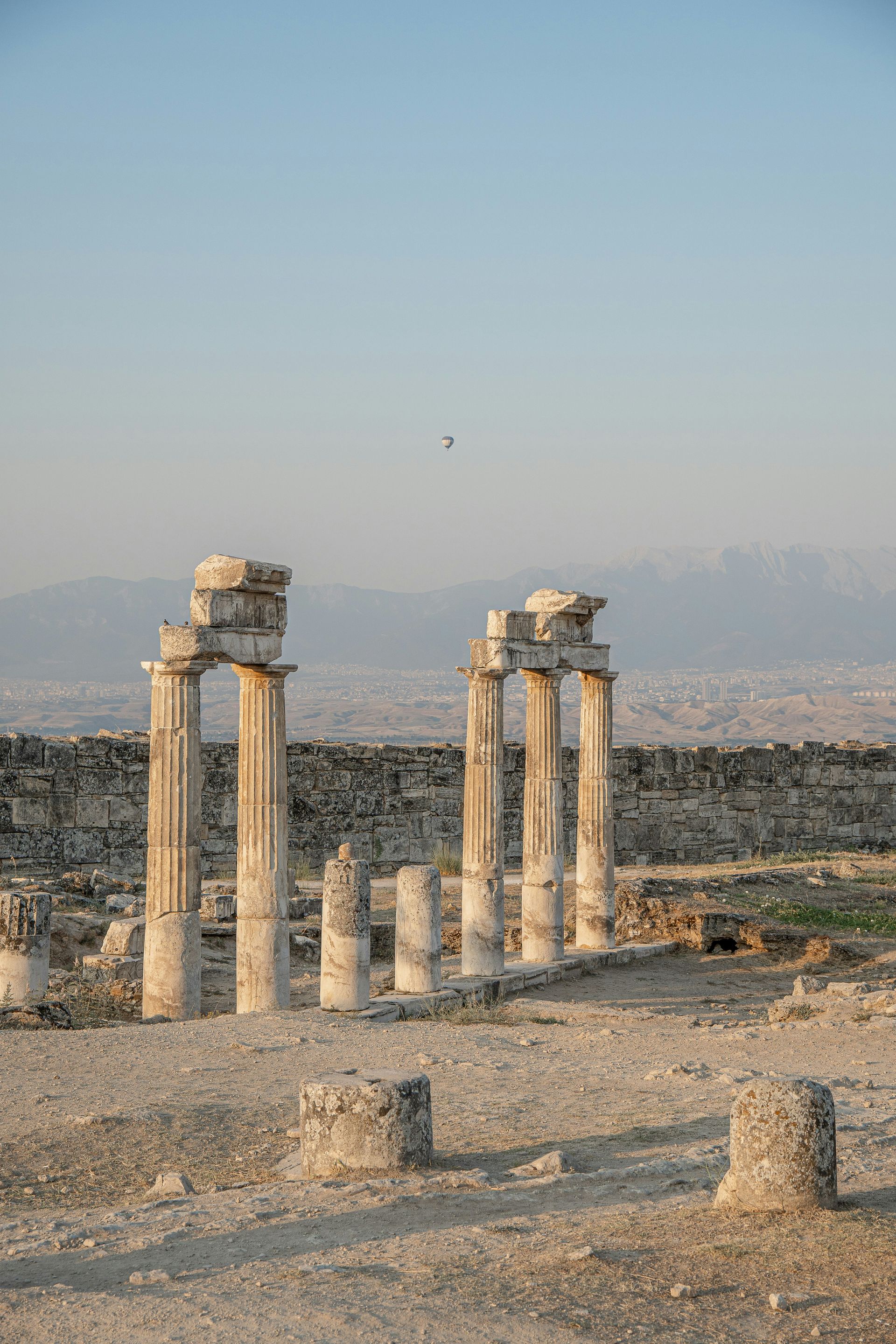 Ruined stone columns stand in Pamukkale, Turkey, with a distant town and mountains in the background.