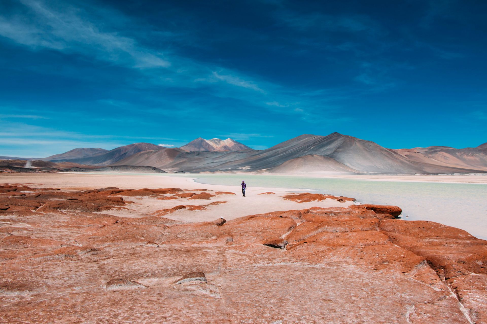 Person standing near a light-colored lake, reddish-brown rock formations, mountains, and a blue sky in the Atacama Desert, Chile.