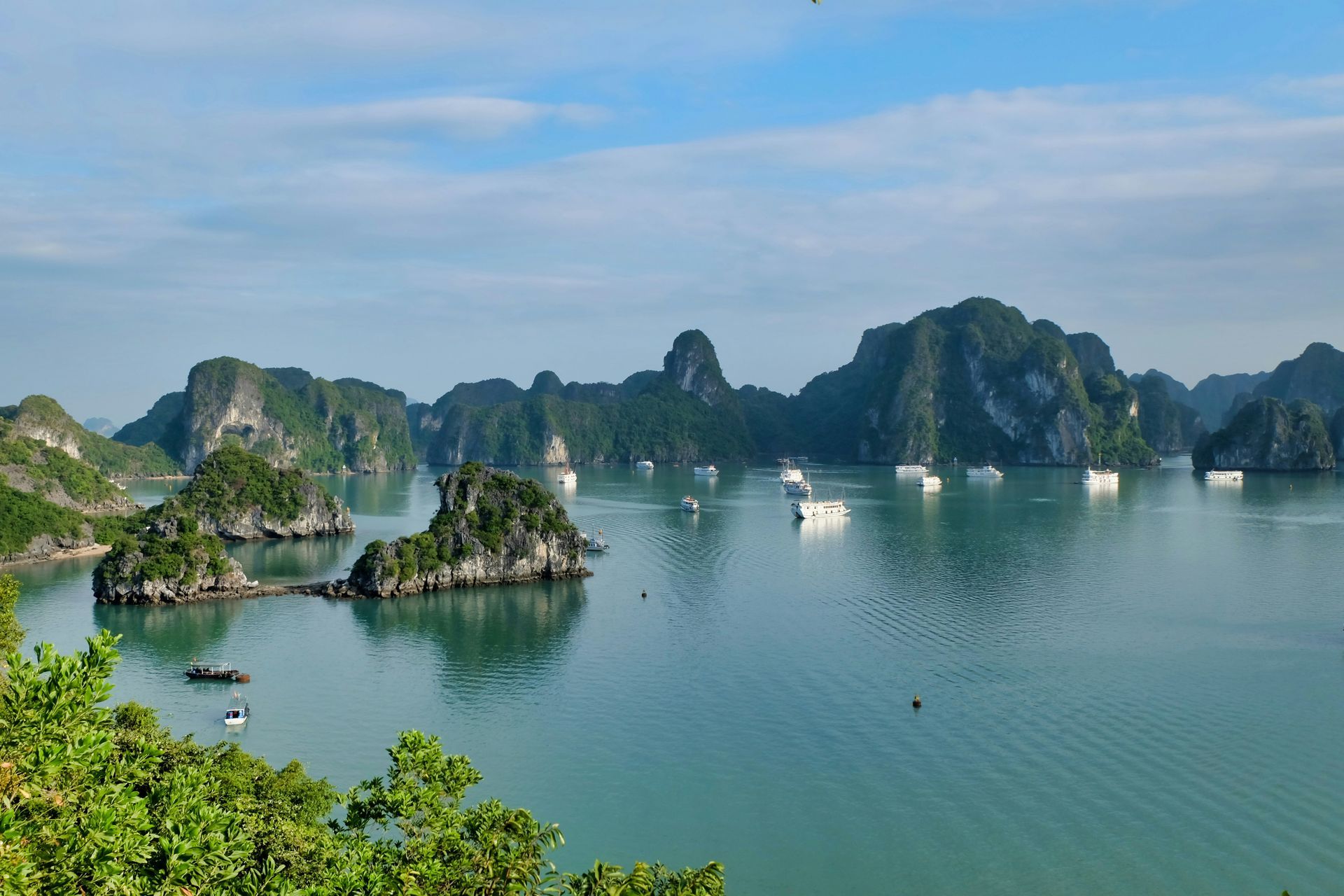 Bay of Halong, Vietnam. Lush green islands rise from calm turquoise water, boats dot the scene under a blue sky.
