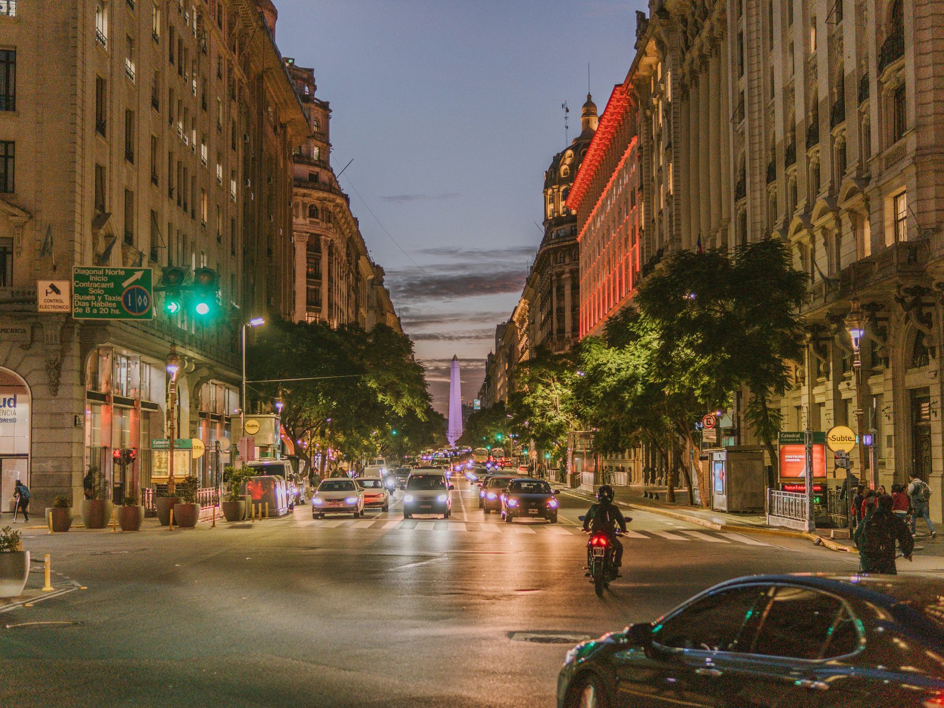 City street at dusk; cars drive towards illuminated obelisk; tall buildings on both sides in Buenos Aires, Argentina.