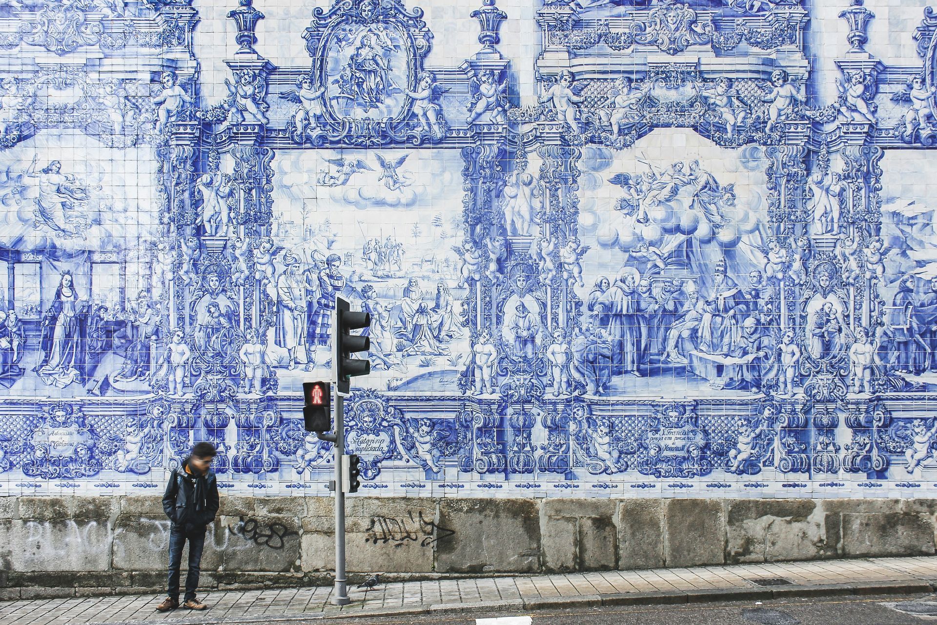 Person standing near traffic light, in front of a wall covered with blue and white azulejos tile artwork in Porto, Portugal.
