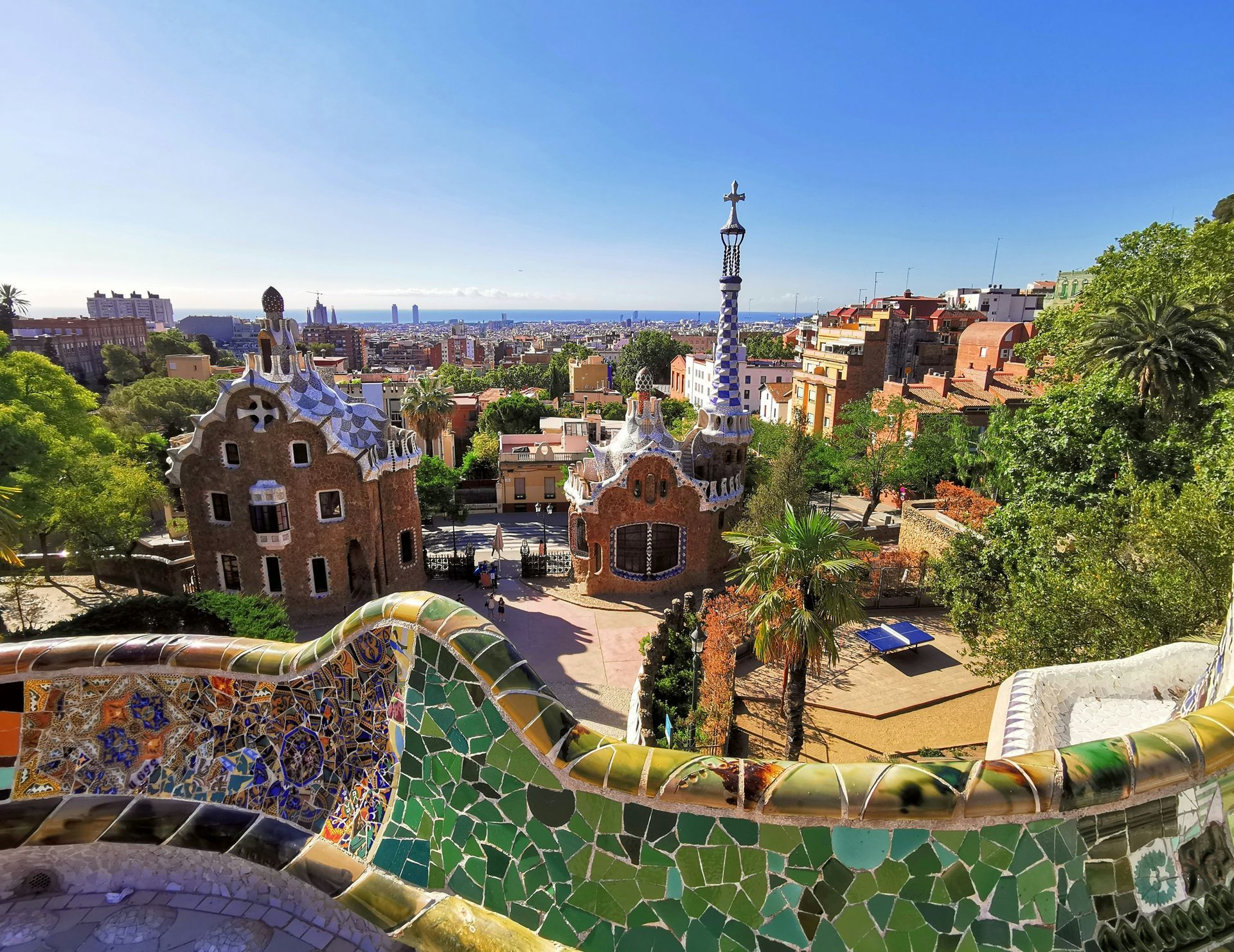 Park Güell, Barcelona. Colorful mosaic benches overlook whimsical buildings, city skyline, and blue sky.