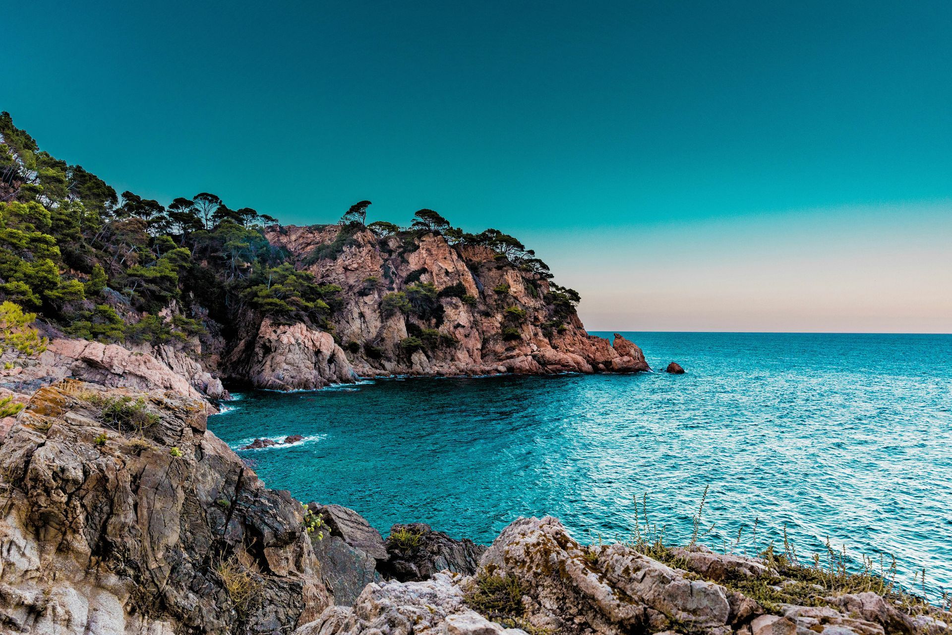 Rocky coastline with turquoise water, a reddish-brown cliff, and a blue sky in Marbella, Spain.