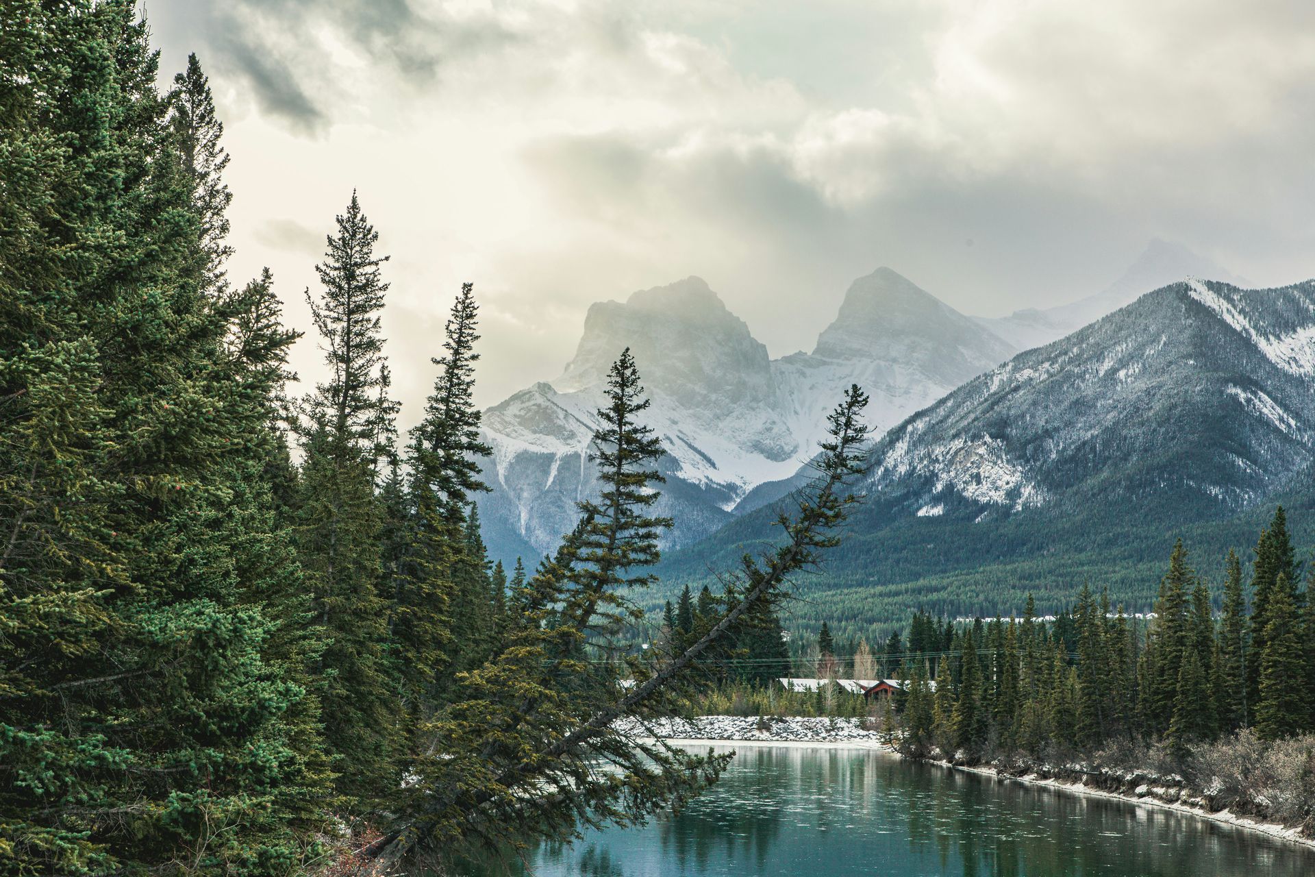 Snowy mountains loom behind a forest of evergreen trees, reflecting in the tranquil river below in Canmore, Canada.