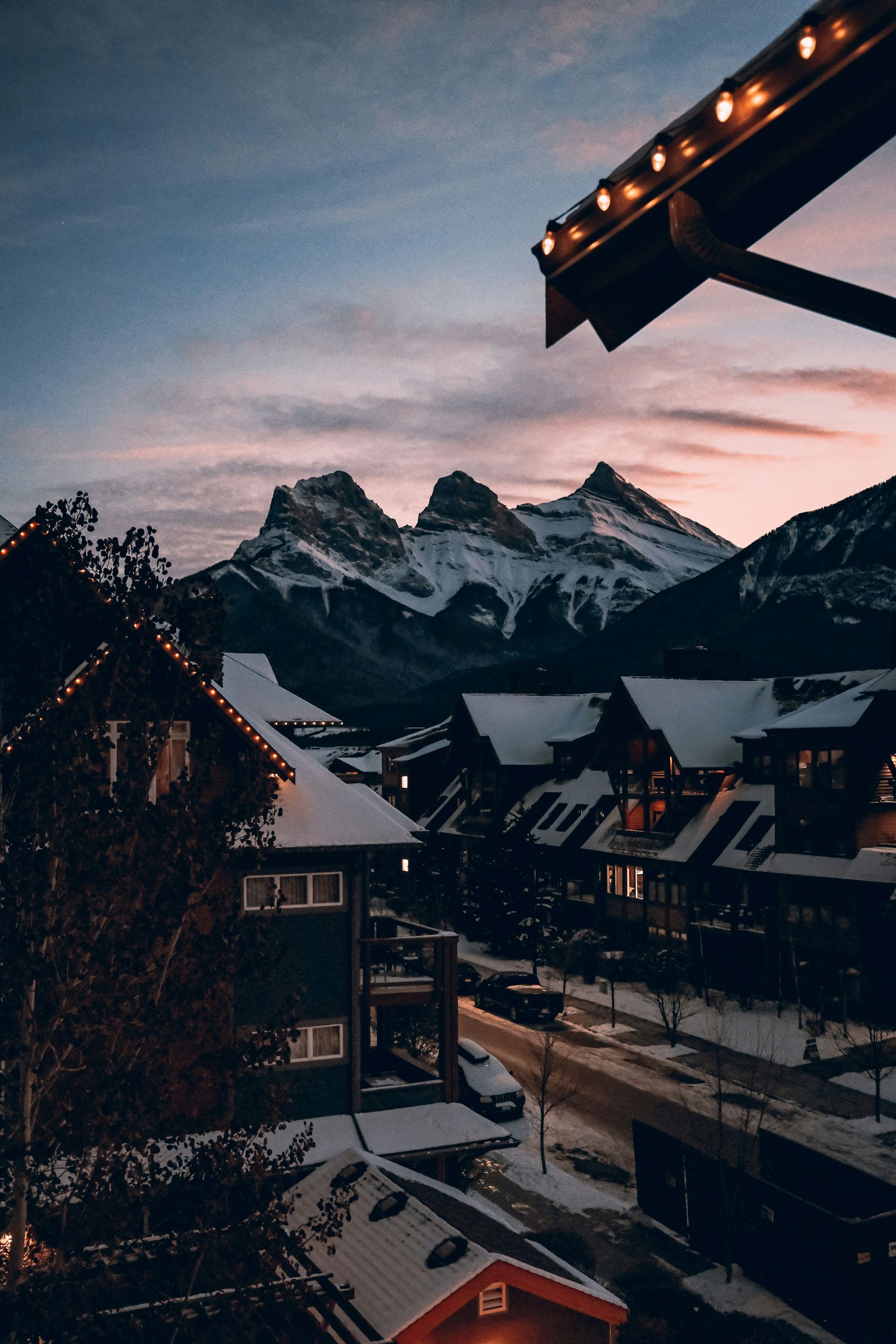 Snow-covered buildings and street at dusk with mountain range in the background. String lights adorn rooftops in Canmore, Canada.