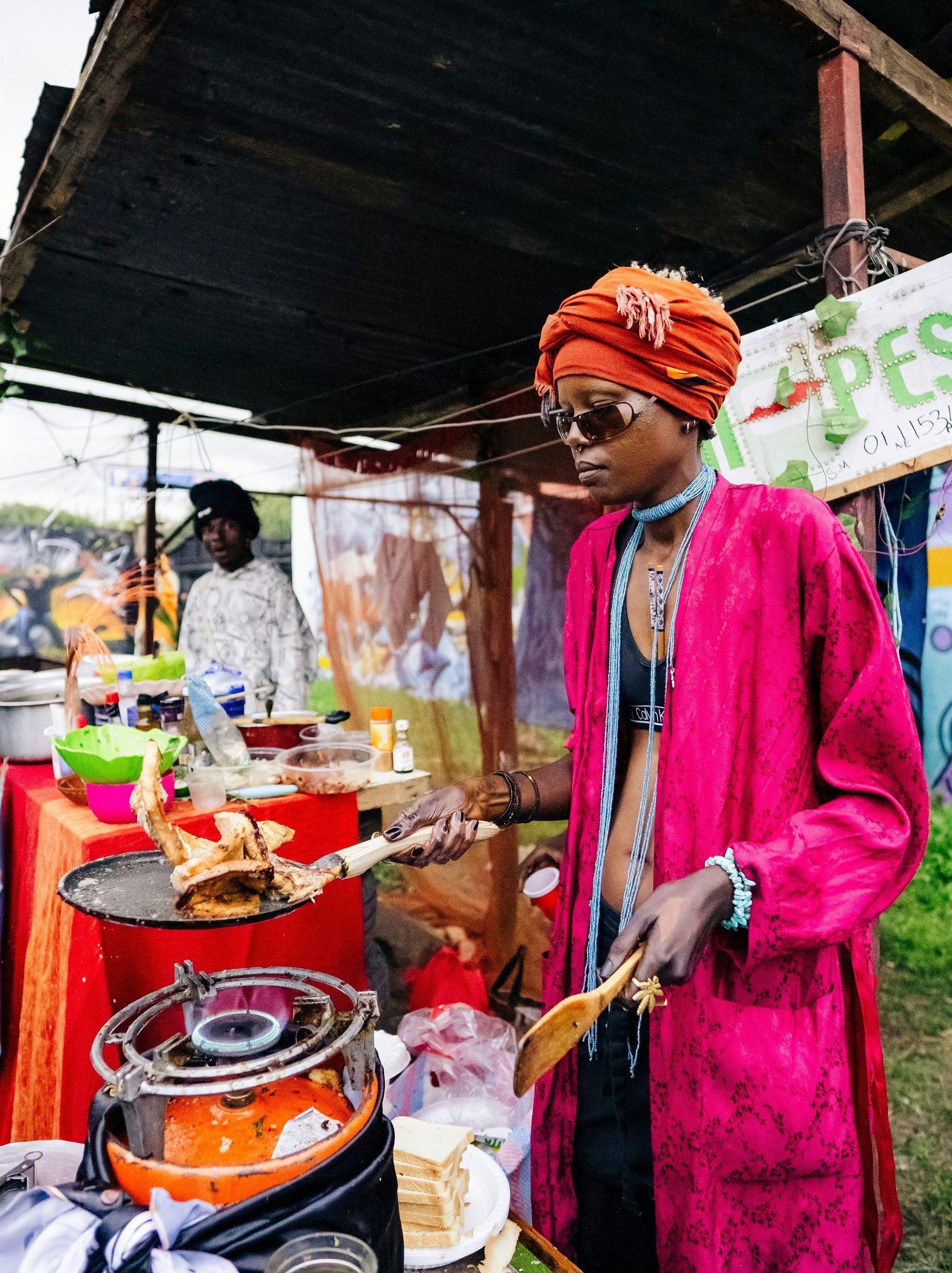 Person in pink robe and turban cooks food at a market stall in Kenya.