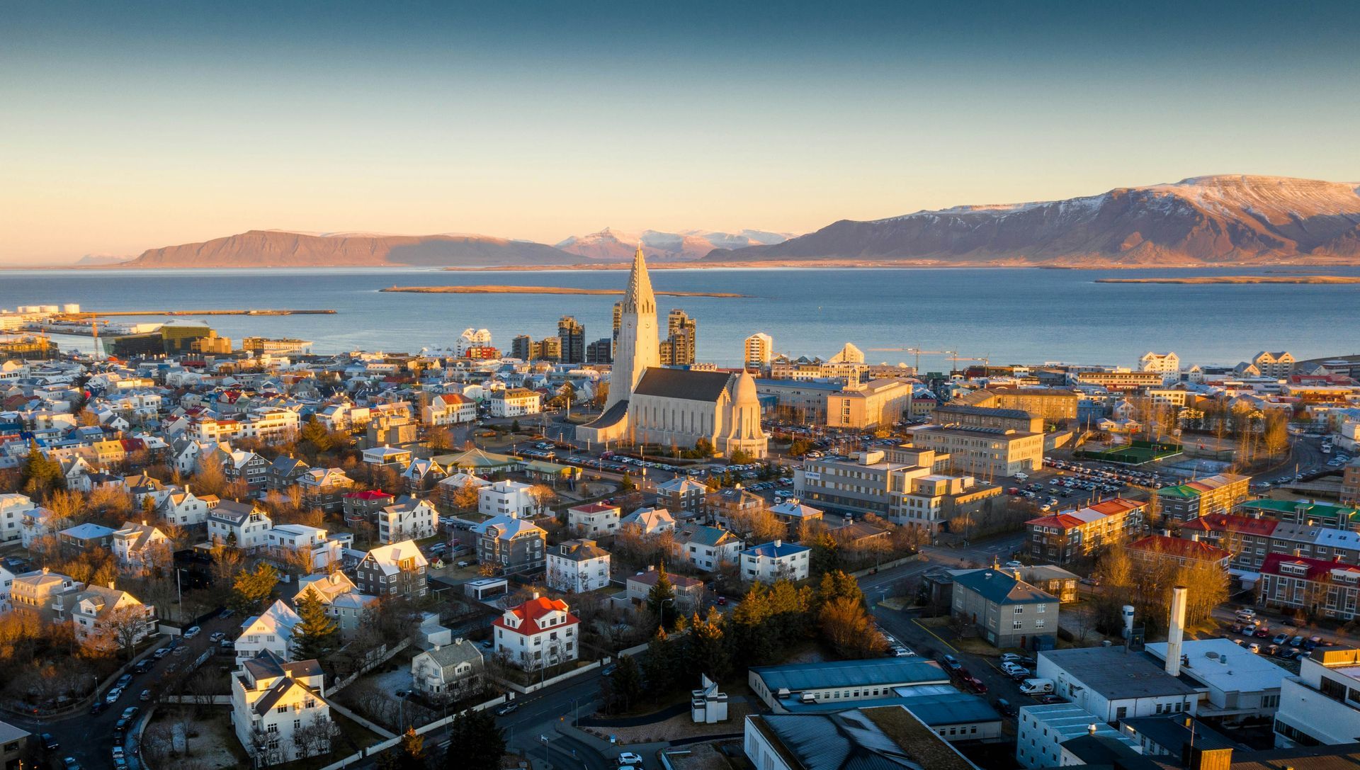 Reykjavik cityscape with church spire, colorful buildings, mountains, and ocean under blue sky.