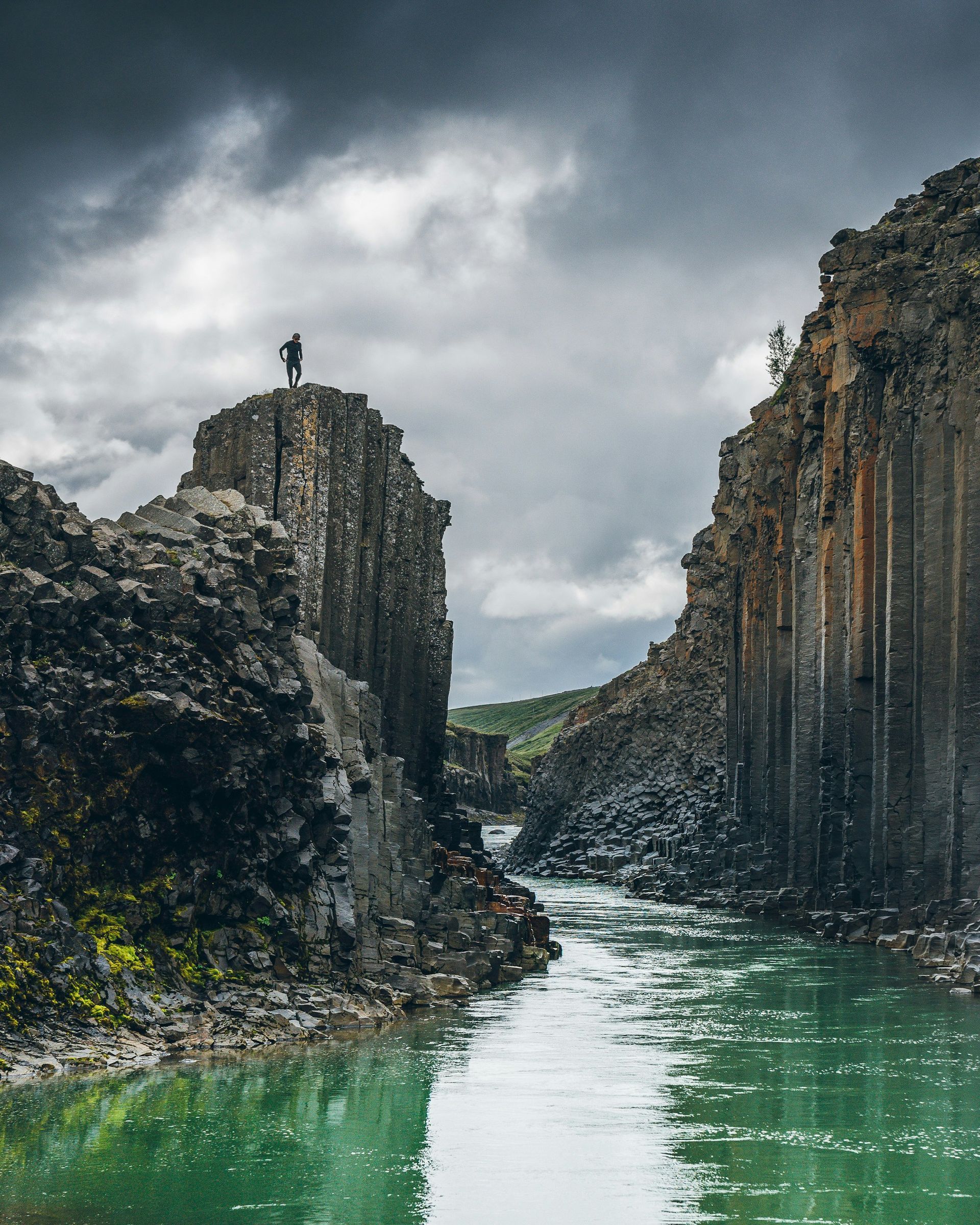 A person stands atop a rock formation, overlooking a river within a basalt Stuðlagil Canyon, Iceland. 
