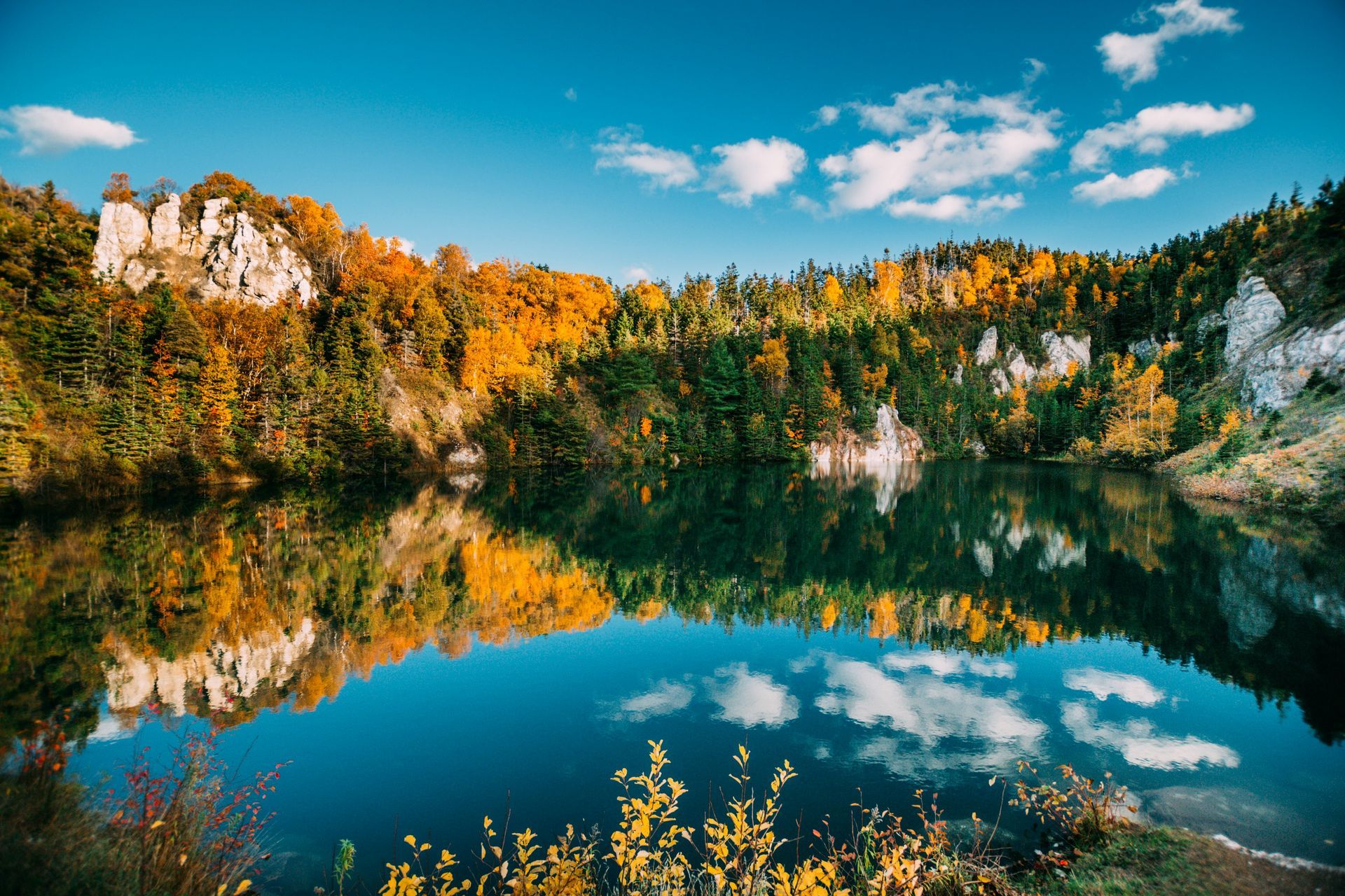 A tranquil lake reflects a vibrant autumn landscape of colorful trees and rock formations under a blue sky with fluffy clouds in Nova Scotia, Canada.