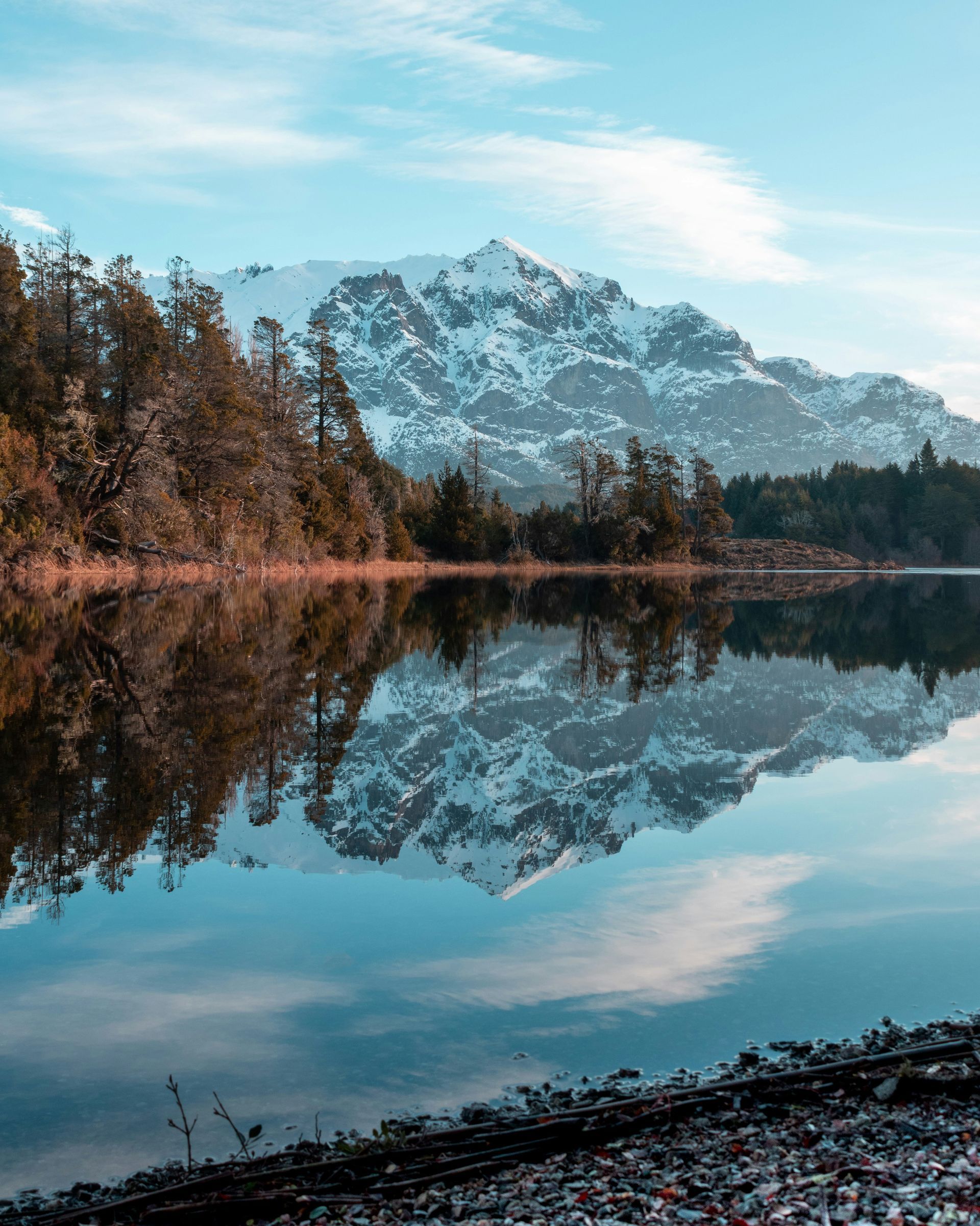 Snow-capped mountain reflecting in still lake, framed by trees and a partly cloudy sky in San Carlos de Bariloche, Argentina.