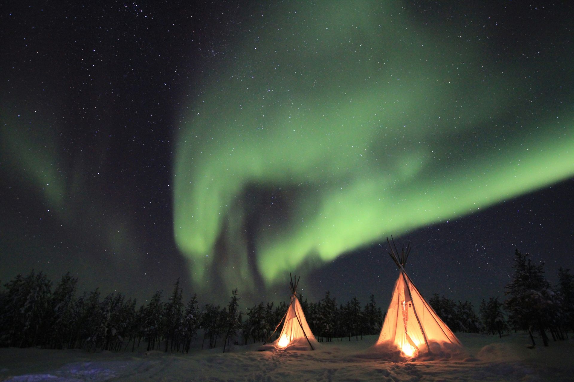 Aurora borealis (Northern Lights) illuminate the night sky above illuminated teepees in a snowy landscape in Yellowknife, Canada.