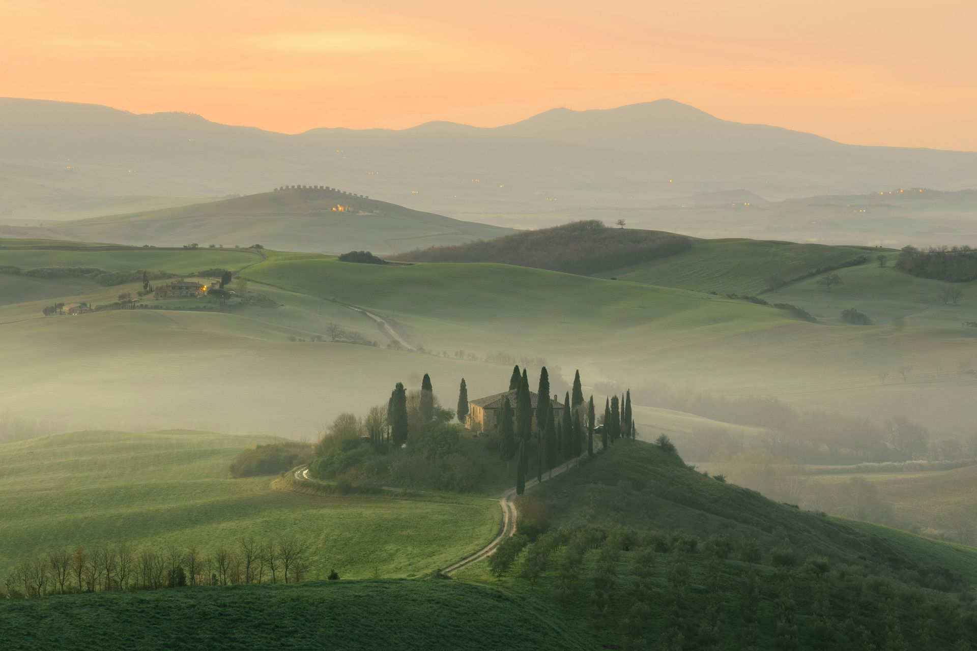 A foggy landscape with trees on a hill in the foreground and mountains in the background in Asciano, Tuscany.