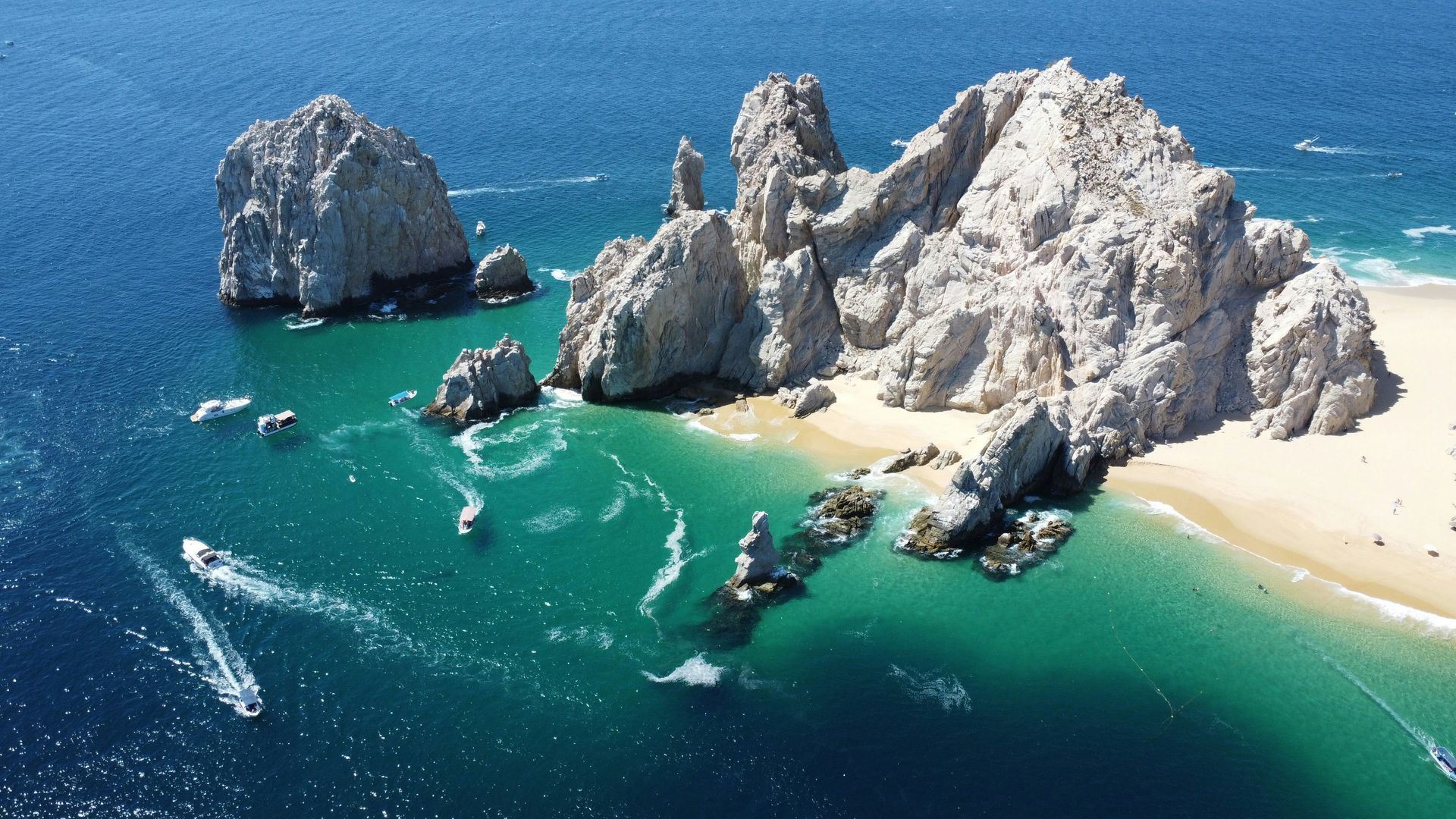 Rocky cliffs jutting from turquoise water, creating a cove filled with boats with a sandy beach Cabo San Lucas, Mexico.