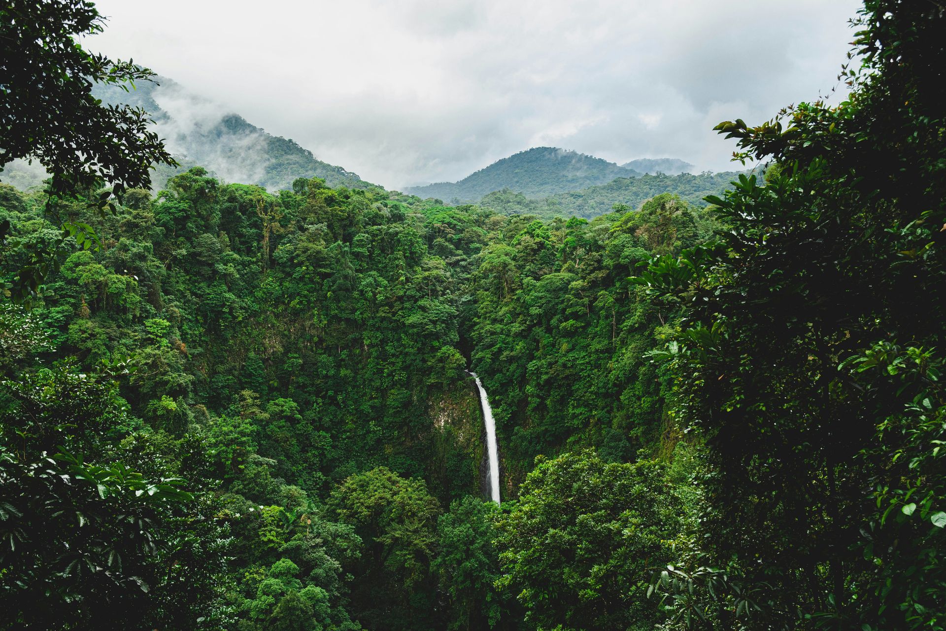 The Fortuna Waterfall cascading through lush green jungle, mountains in background under a cloudy sky in Costa Rica.