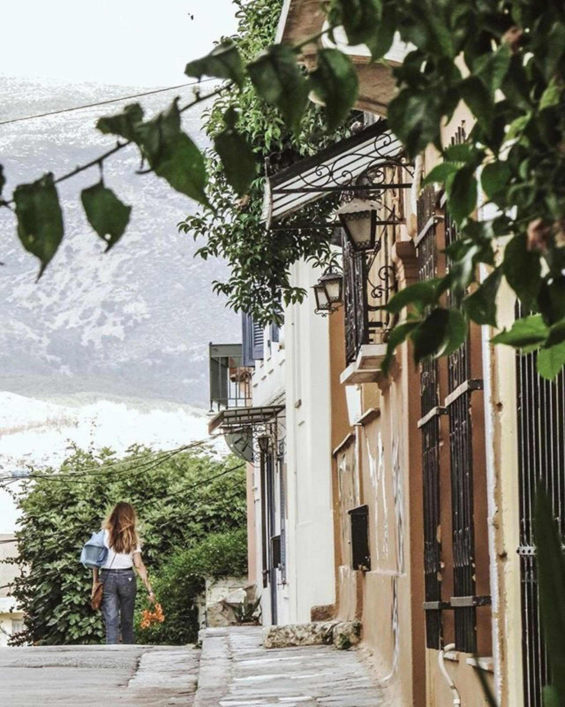 A woman is walking down a narrow street with mountains in the background in Athens, Greece.