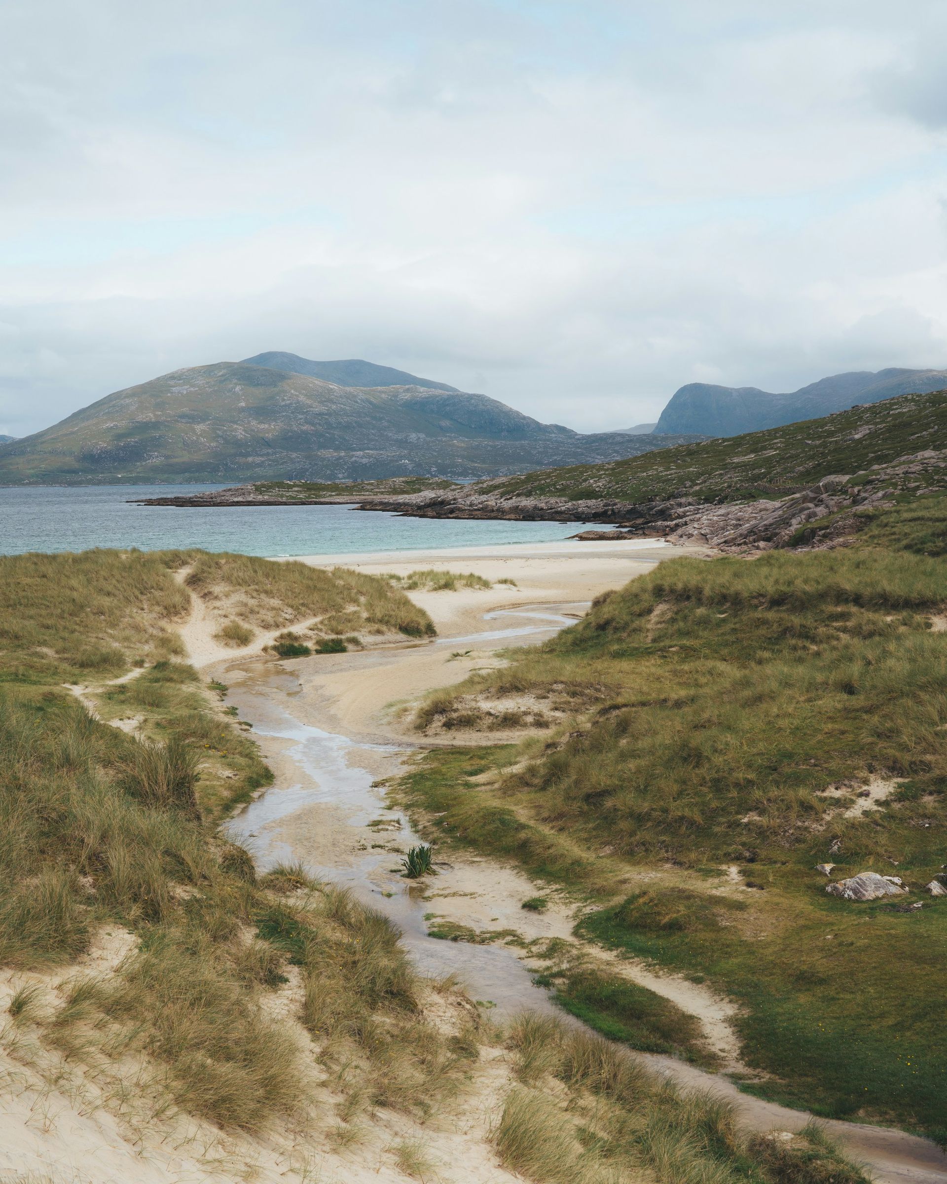 A beach with a stream flowing through grassy dunes, mountains in the background on the Hebrides Islands, Scotland.