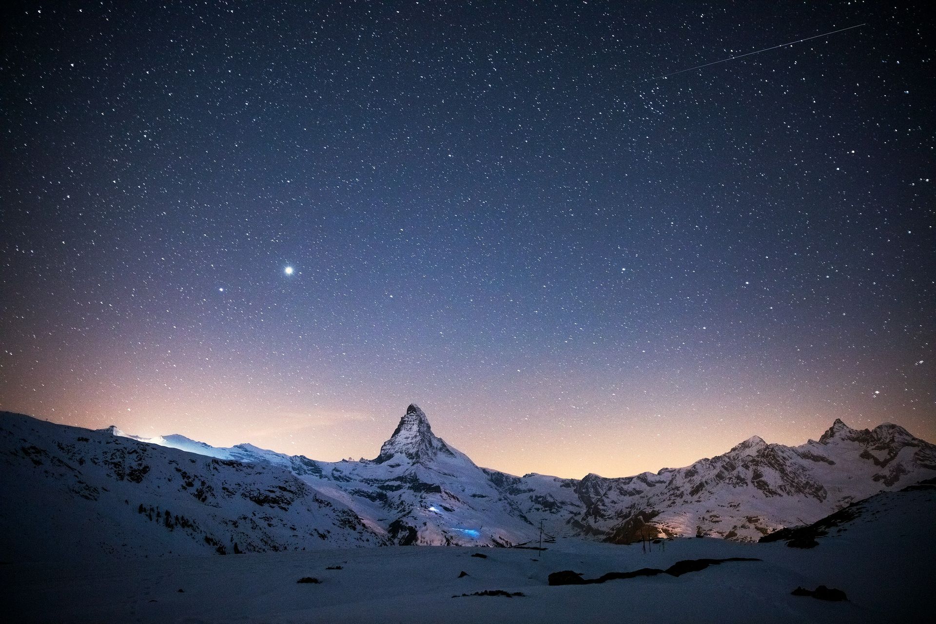 Snowy mountain range under a dark blue starry night sky with the Matterhorn peak illuminated by a faint glow.