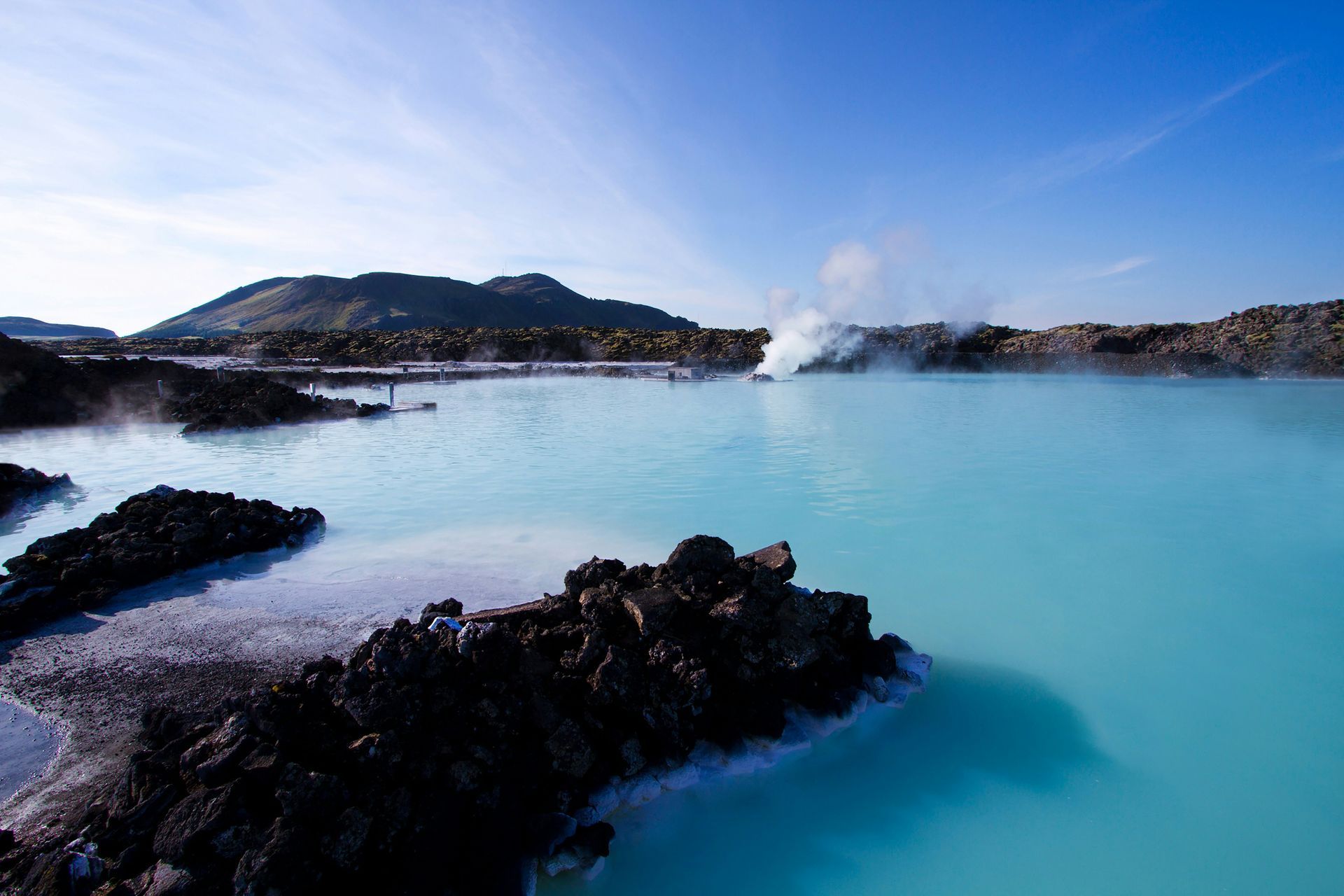 A turquoise geothermal lagoon in Iceland with dark volcanic rocks, steam rising, and a mountain in the background.