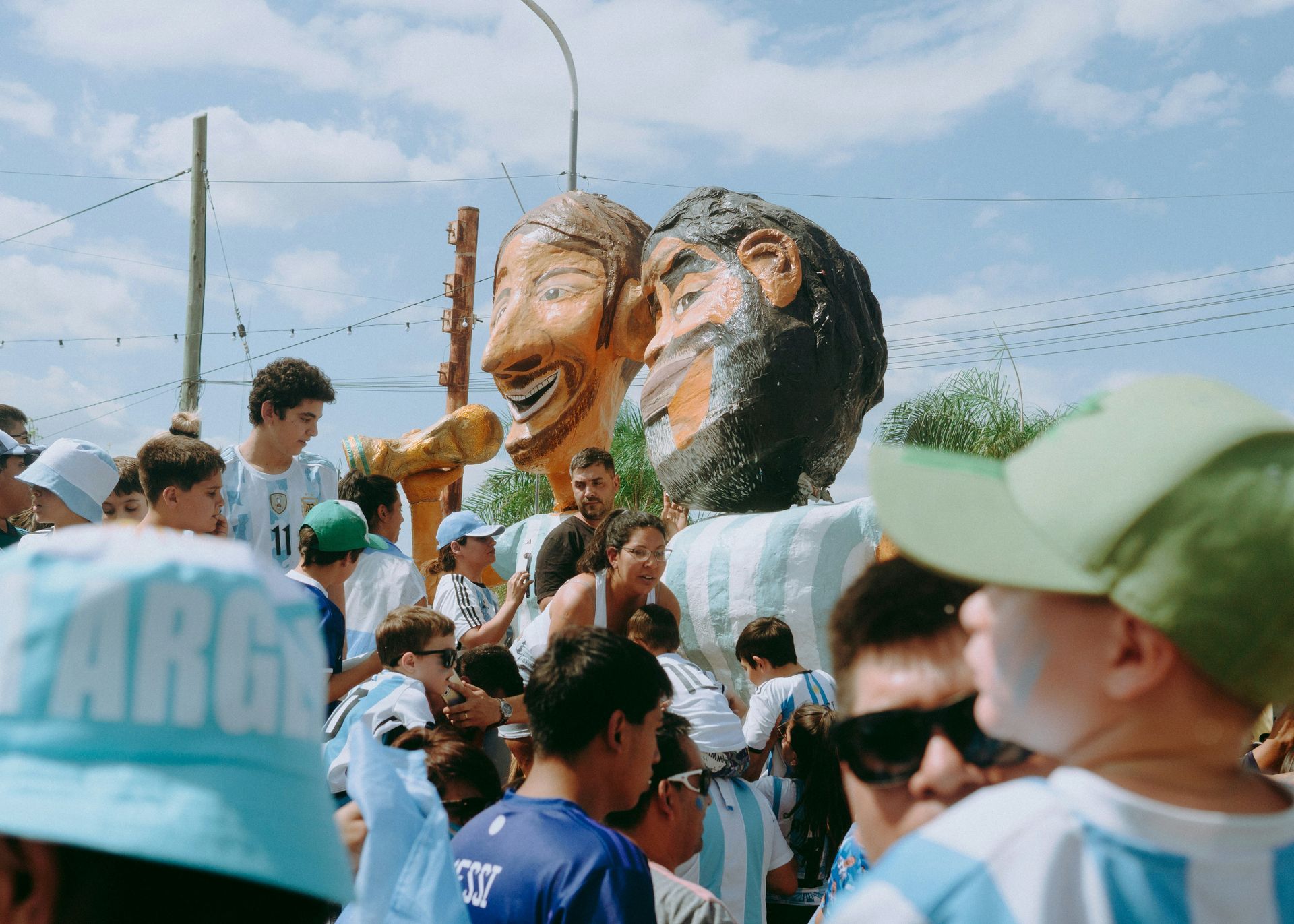 Crowd celebrates, giant heads of Maradona & soccer fans smiling, blue & white, sunny day in Argentina. 