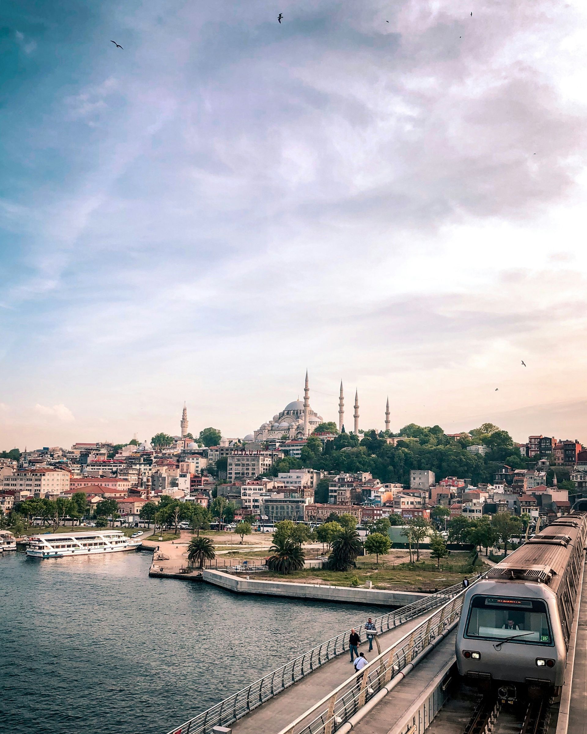 Train crossing a bridge in Istanbul with cityscape, water, and mosque under a cloudy sky.