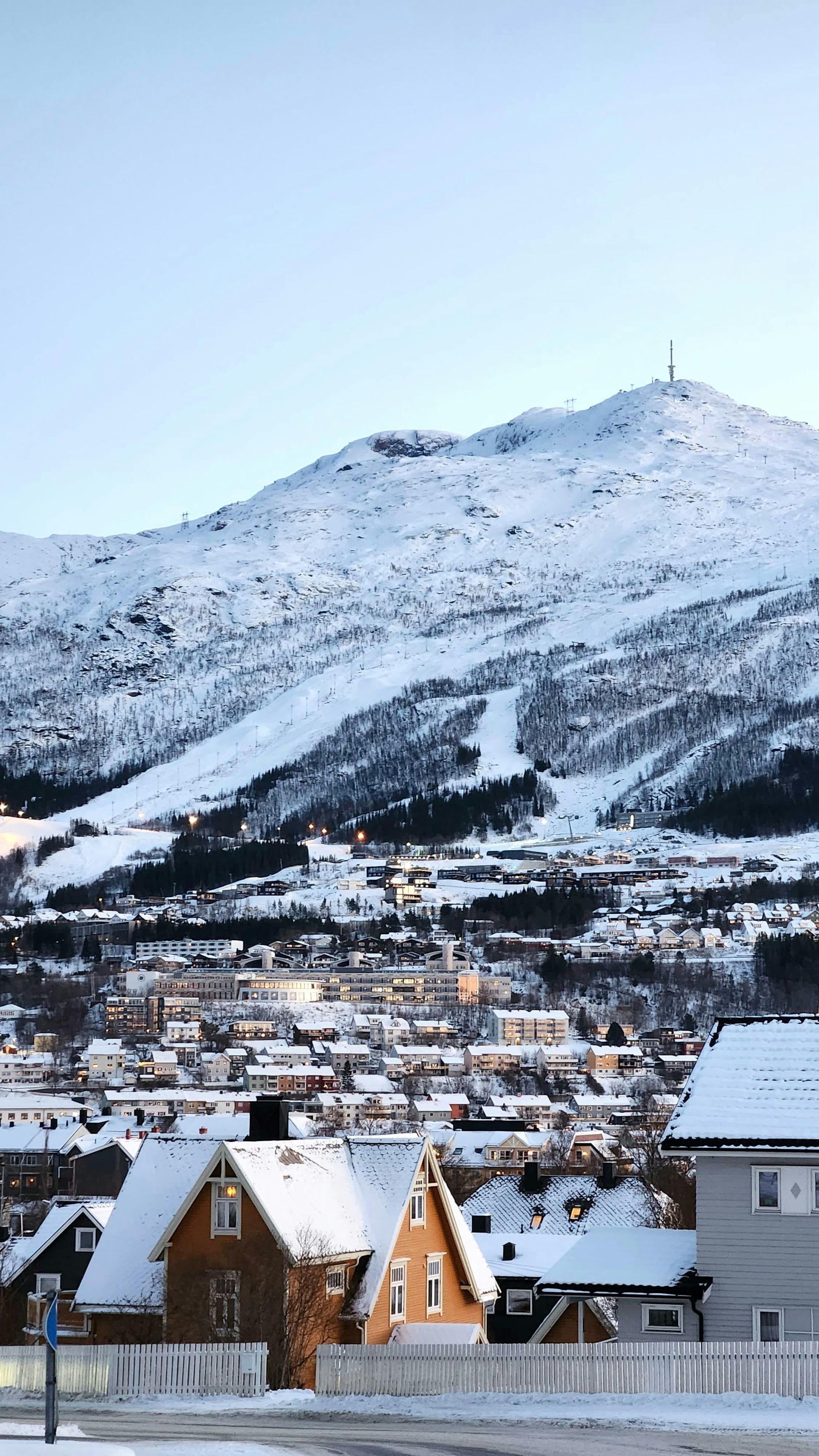 Snow-covered town at the base of a mountain with a ski slope, under a clear, light-blue sky in Narvik, Norway.