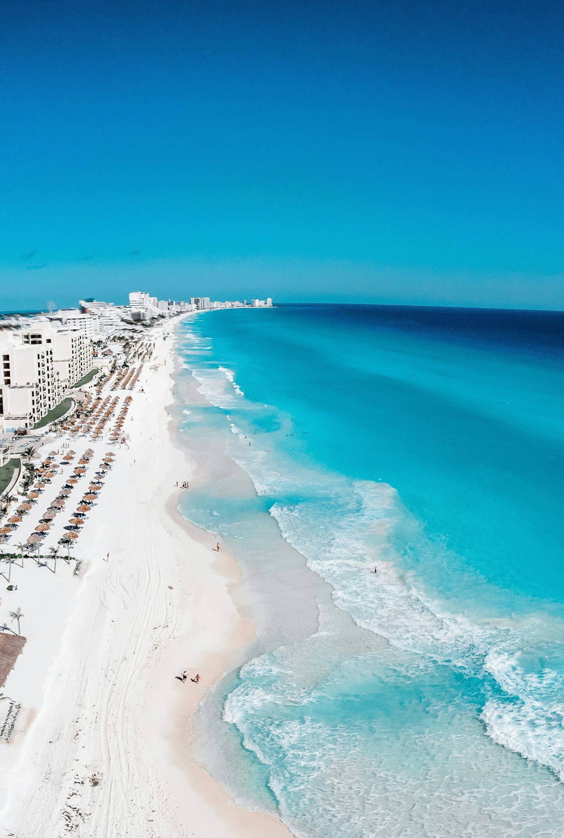 Aerial view of a white sandy beach with turquoise water and buildings under a bright blue sky in Cancún, Mexico.