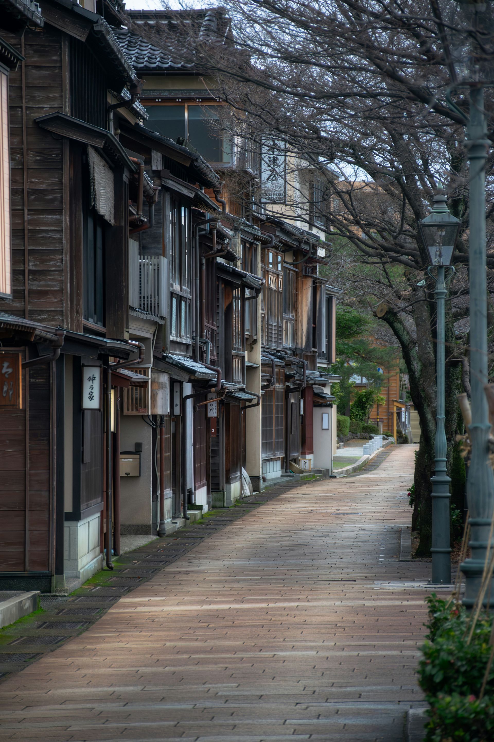 Narrow cobblestone street lined with aged wooden buildings and bare trees in Kanazawa, Japan.
