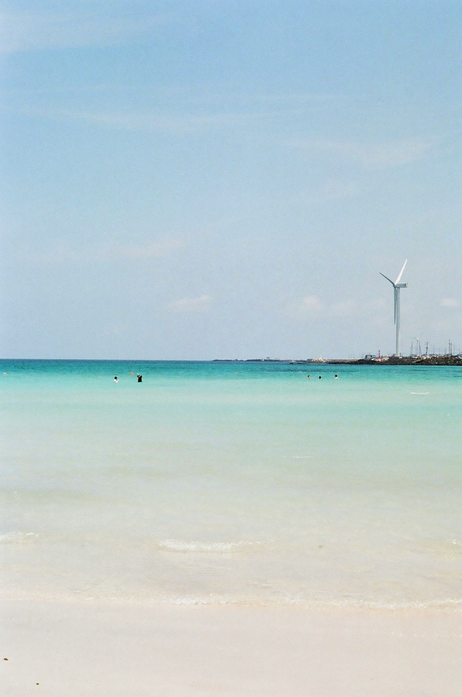 Beach scene with turquoise water, white sand, and a wind turbine on the horizon on Jeju Island, South Korea.