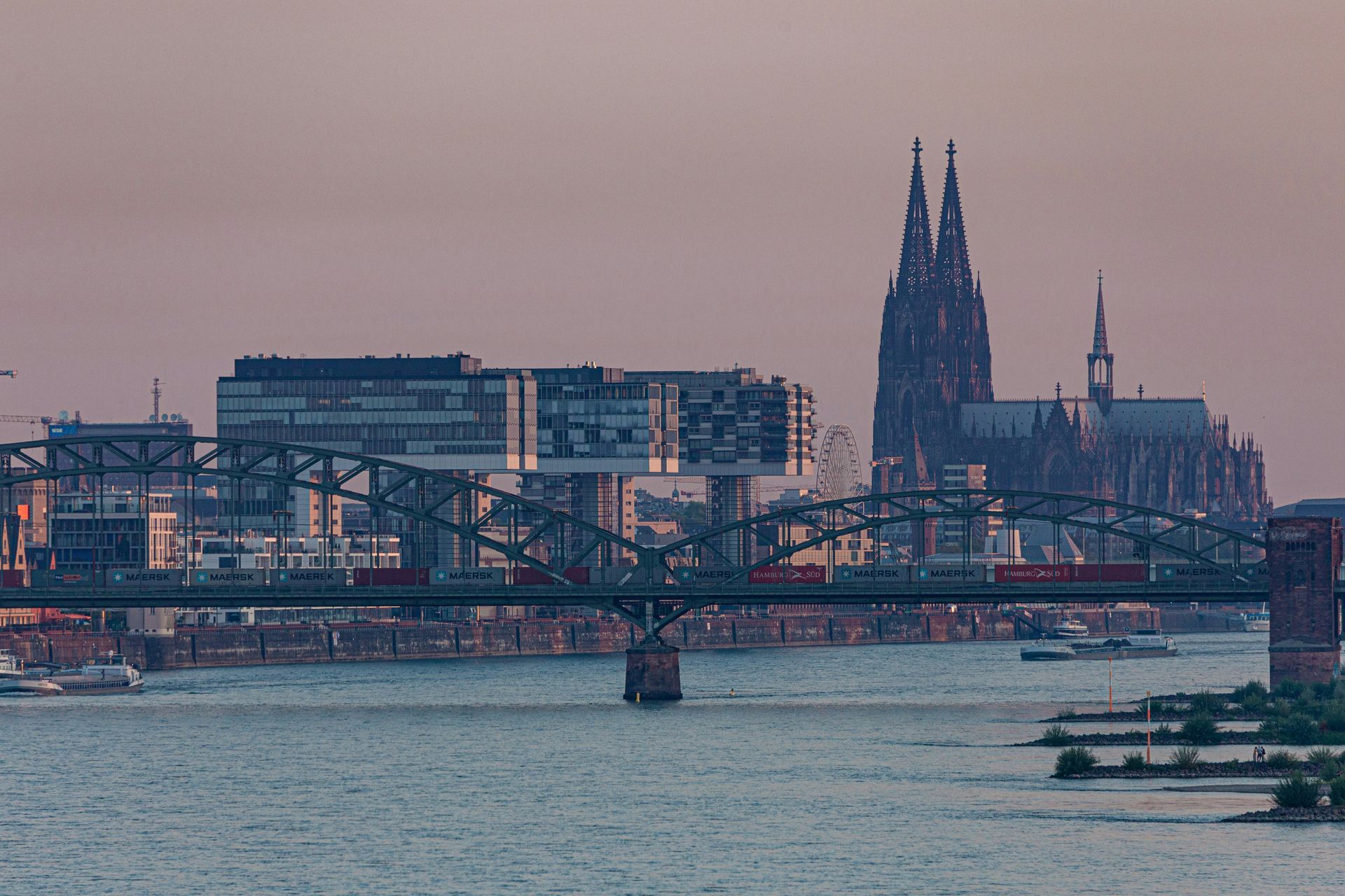 Cologne cityscape with bridge, modern buildings, and Cologne Cathedral against a dusky sky over the Rhine river.