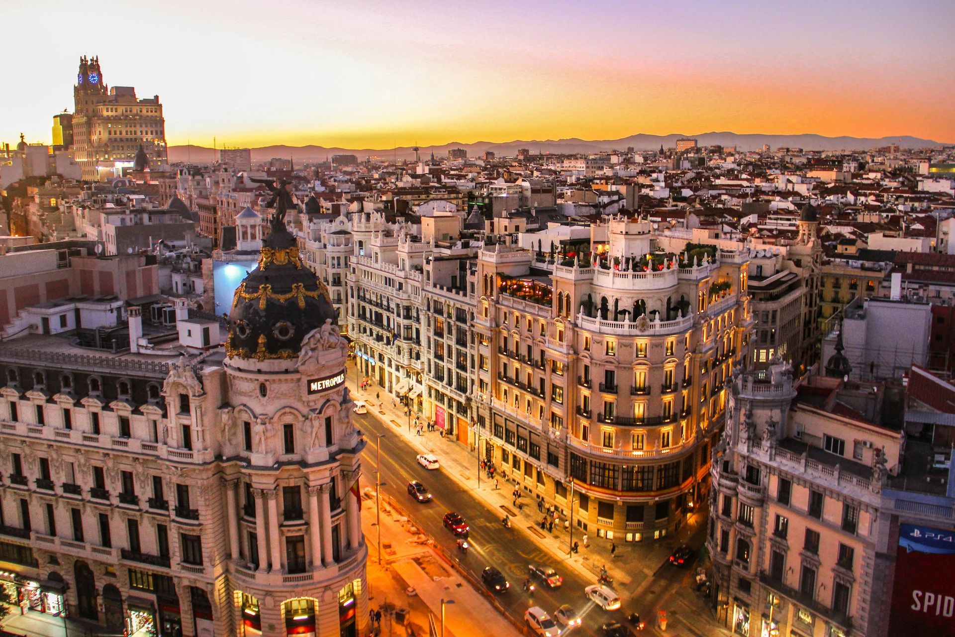 Madrid cityscape at dusk, buildings with illuminated facades, street with traffic, sunset sky.