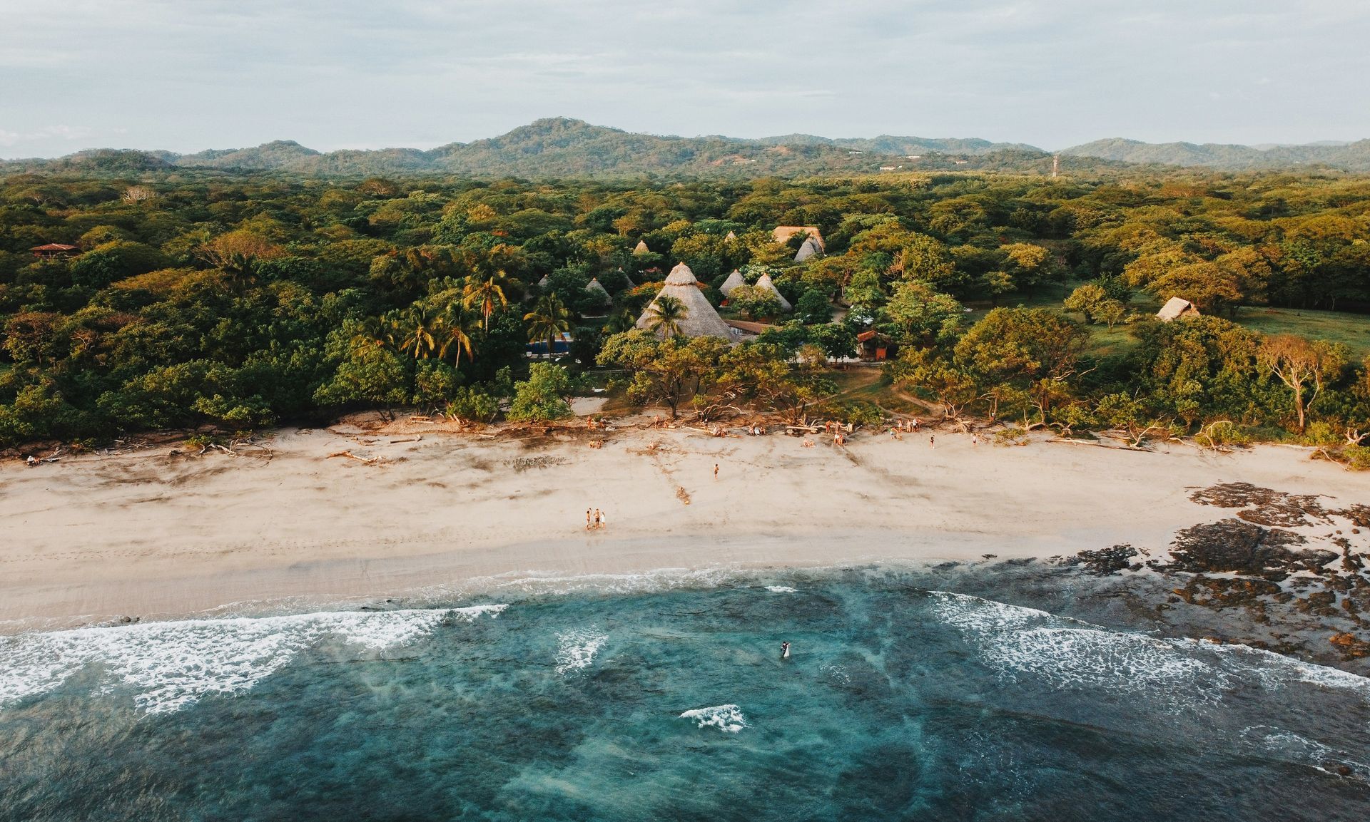 A sandy beach meeting blue-green ocean, with a dense green forest bordering the shore under a cloudy sky in Costa Rica.