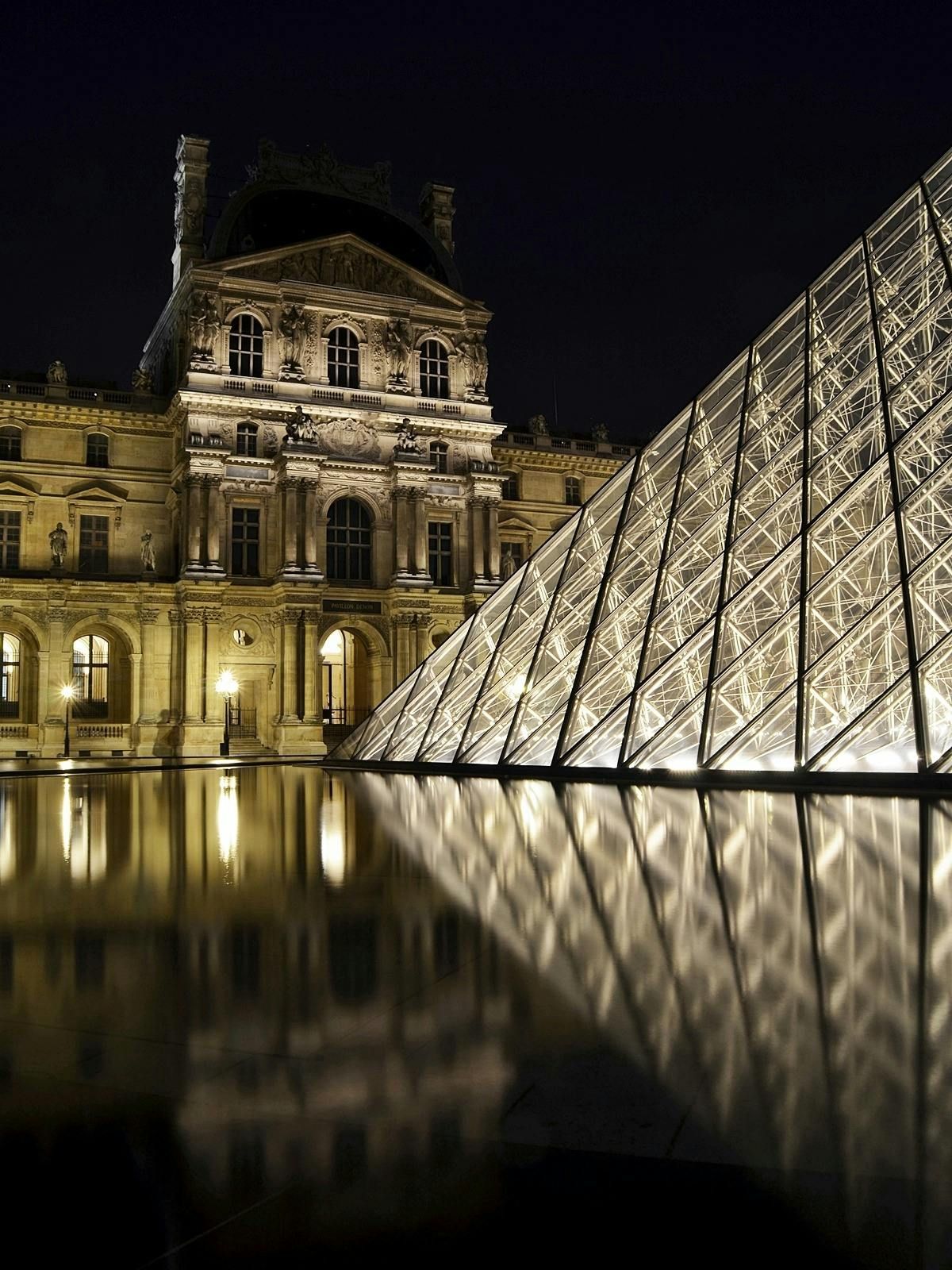 A large building with the Louvre pyramid in front of it is lit up at night in Paris.