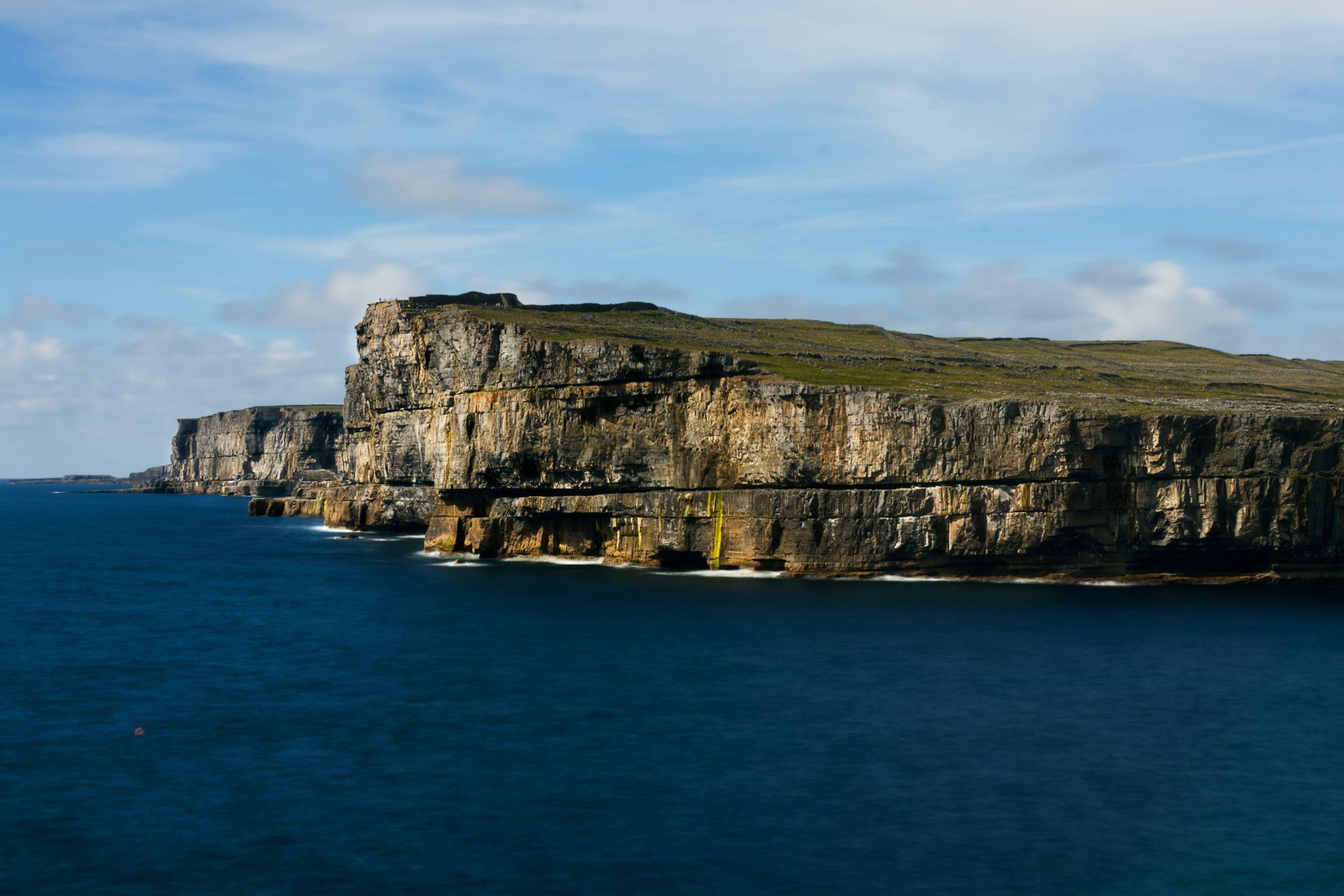 Cliffs rising from the dark blue ocean under a partly cloudy sky on the Aran Islands, Ireland.