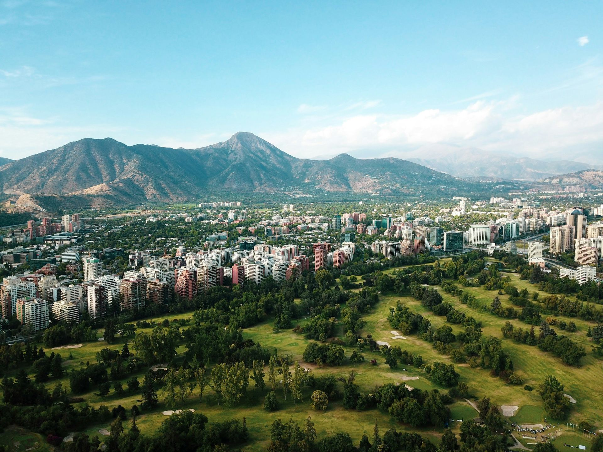 Aerial view of Santiago, Chile, with buildings and green spaces, mountains in the background under a blue sky.