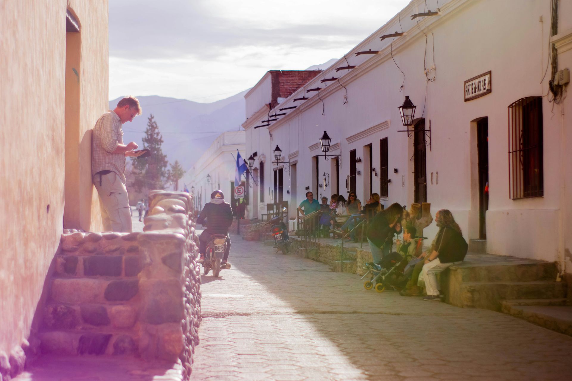 People on sidewalk, motorcyclist, man reading on wall. White buildings, cobblestone street, mountains in Salta, Argentina.