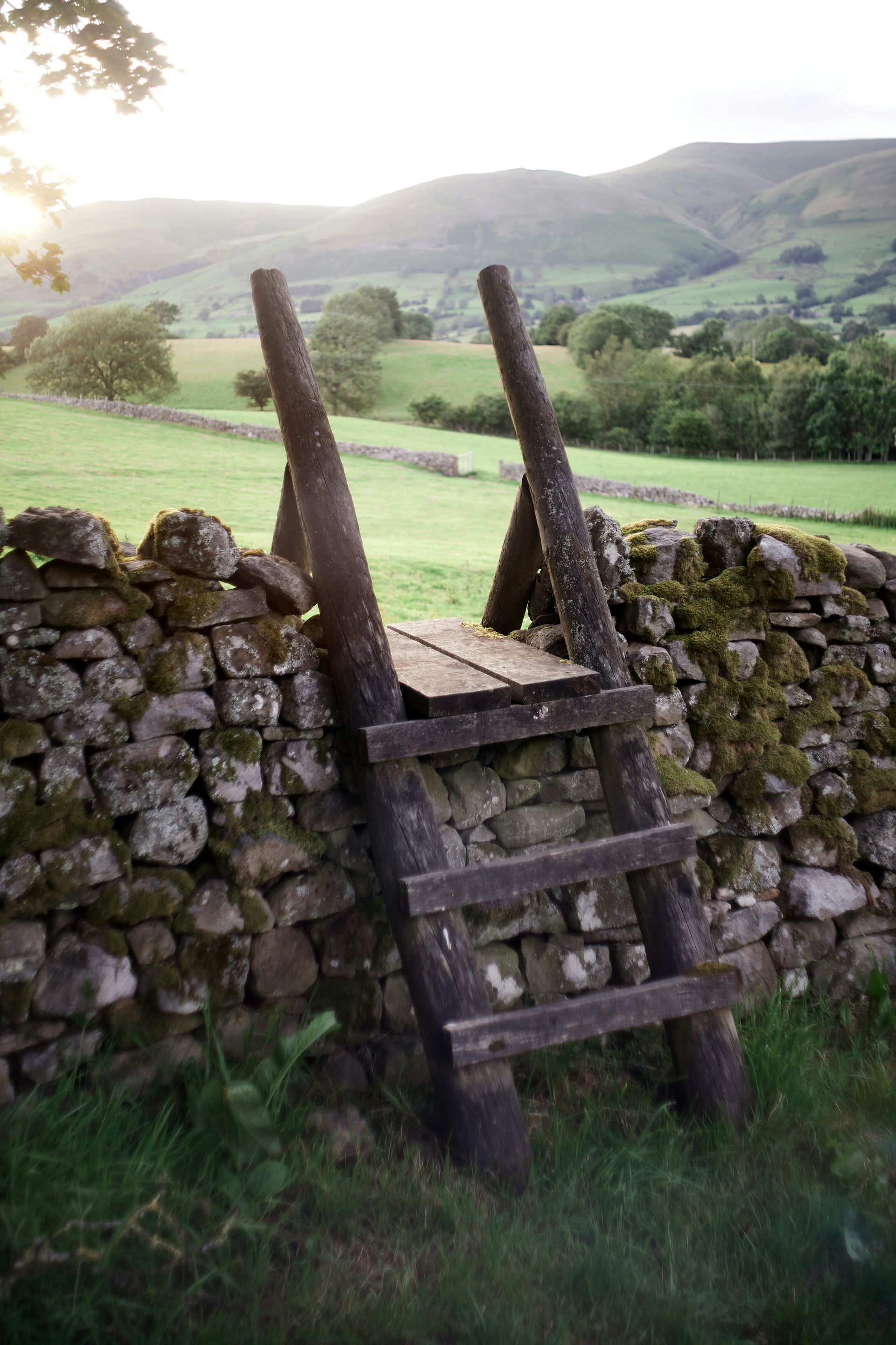Wooden stile over stone wall in a green field with rolling hills in the background in the Yorkshire Dales, England.