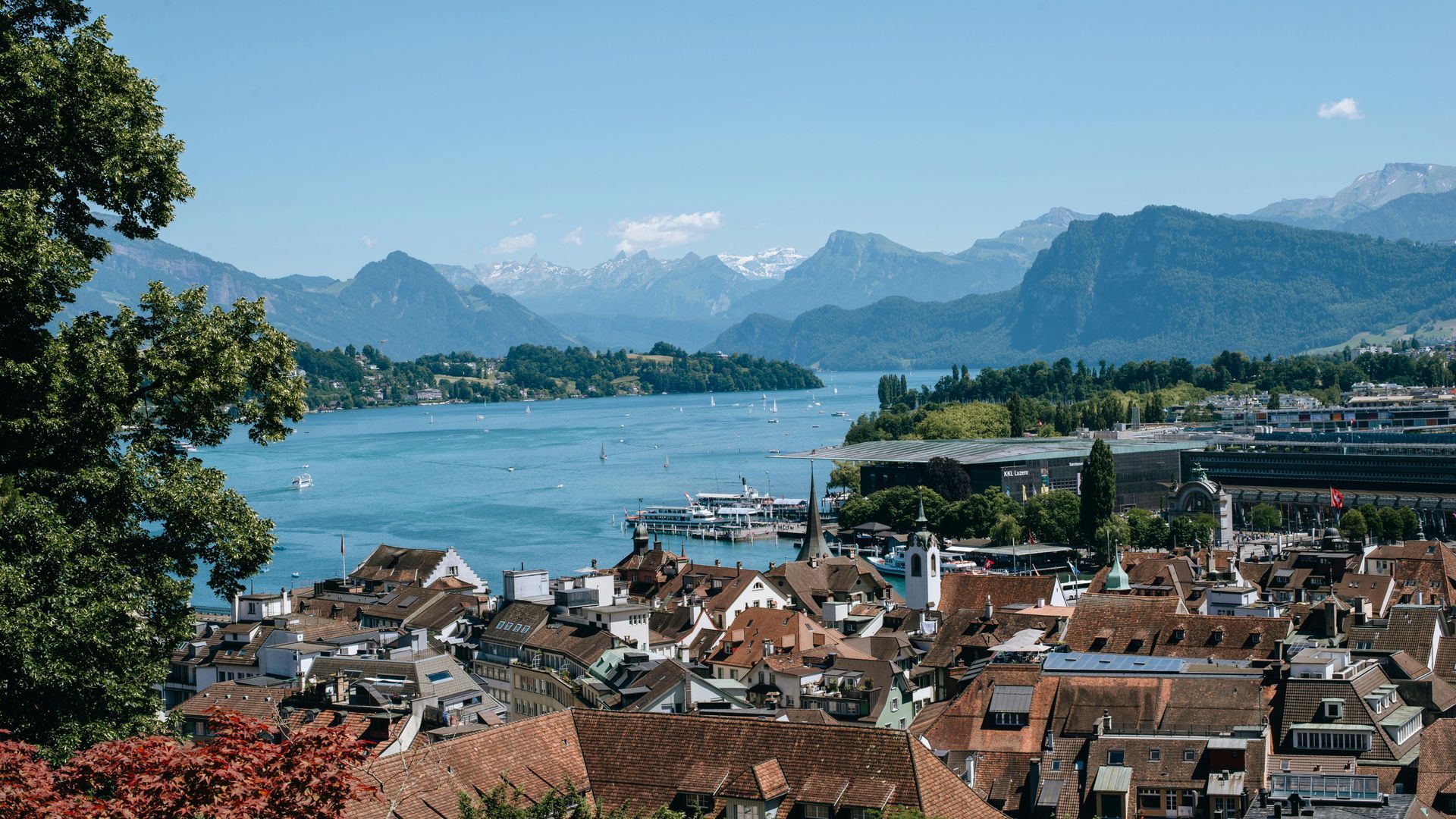 Lucerne, Switzerland overlooking a lake and mountains. Buildings with red roofs, blue water, green trees, and snowy peaks.