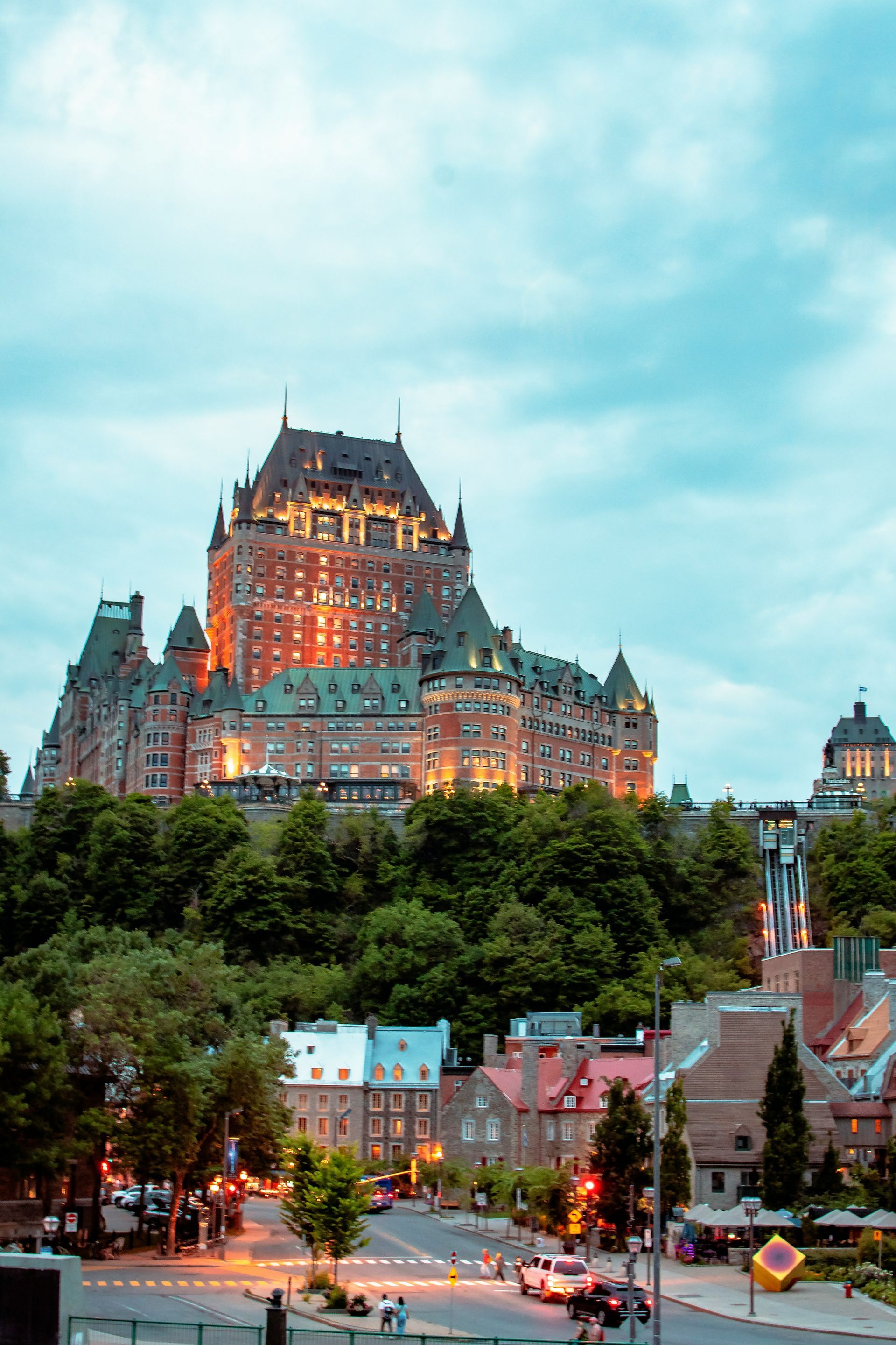 Château Frontenac lit up at dusk, overlooking Quebec City. Buildings and street with trees in foreground.
