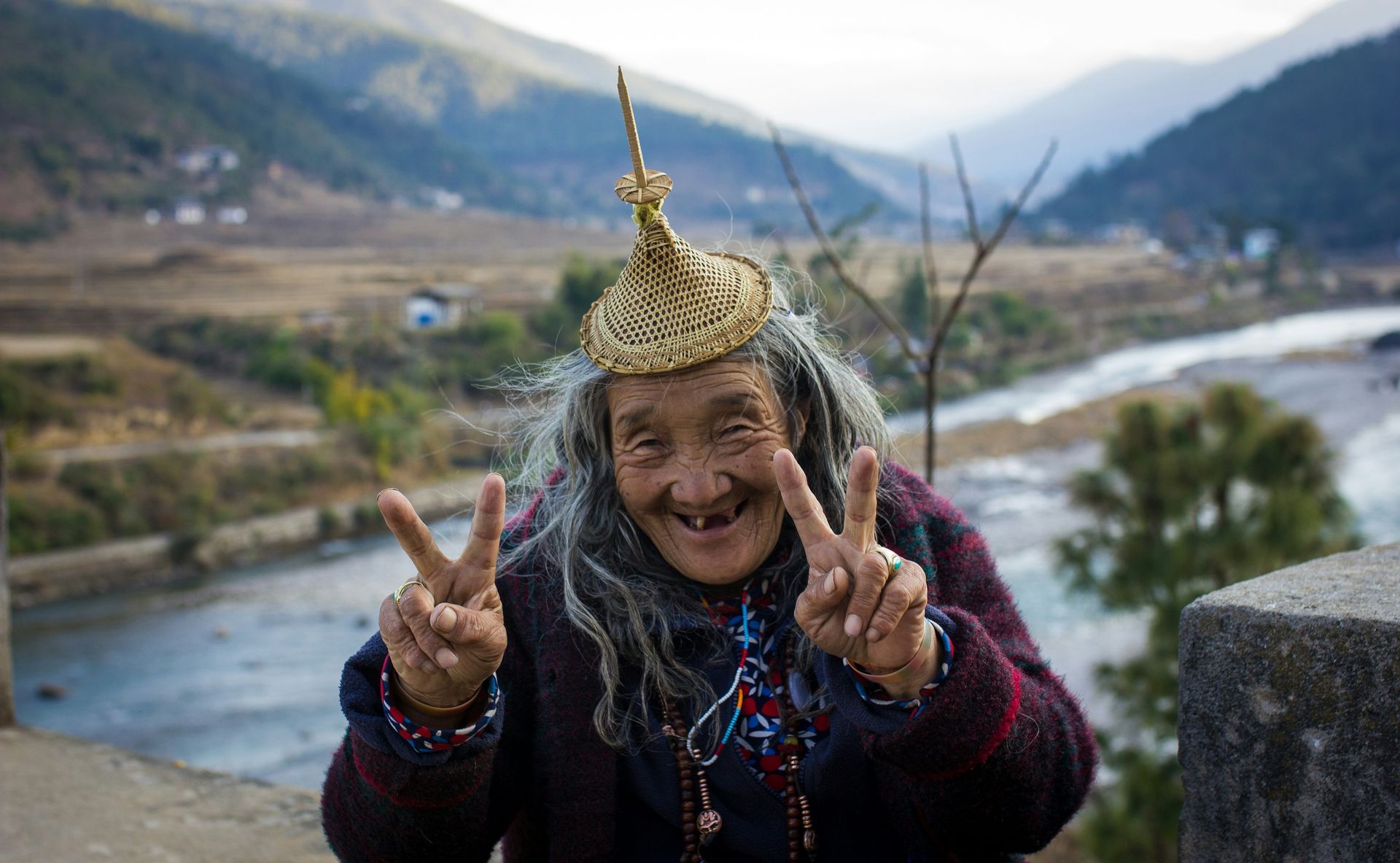 Woman with a decorative gold hat, giving peace signs in Bhutan. Mountainous background, river.