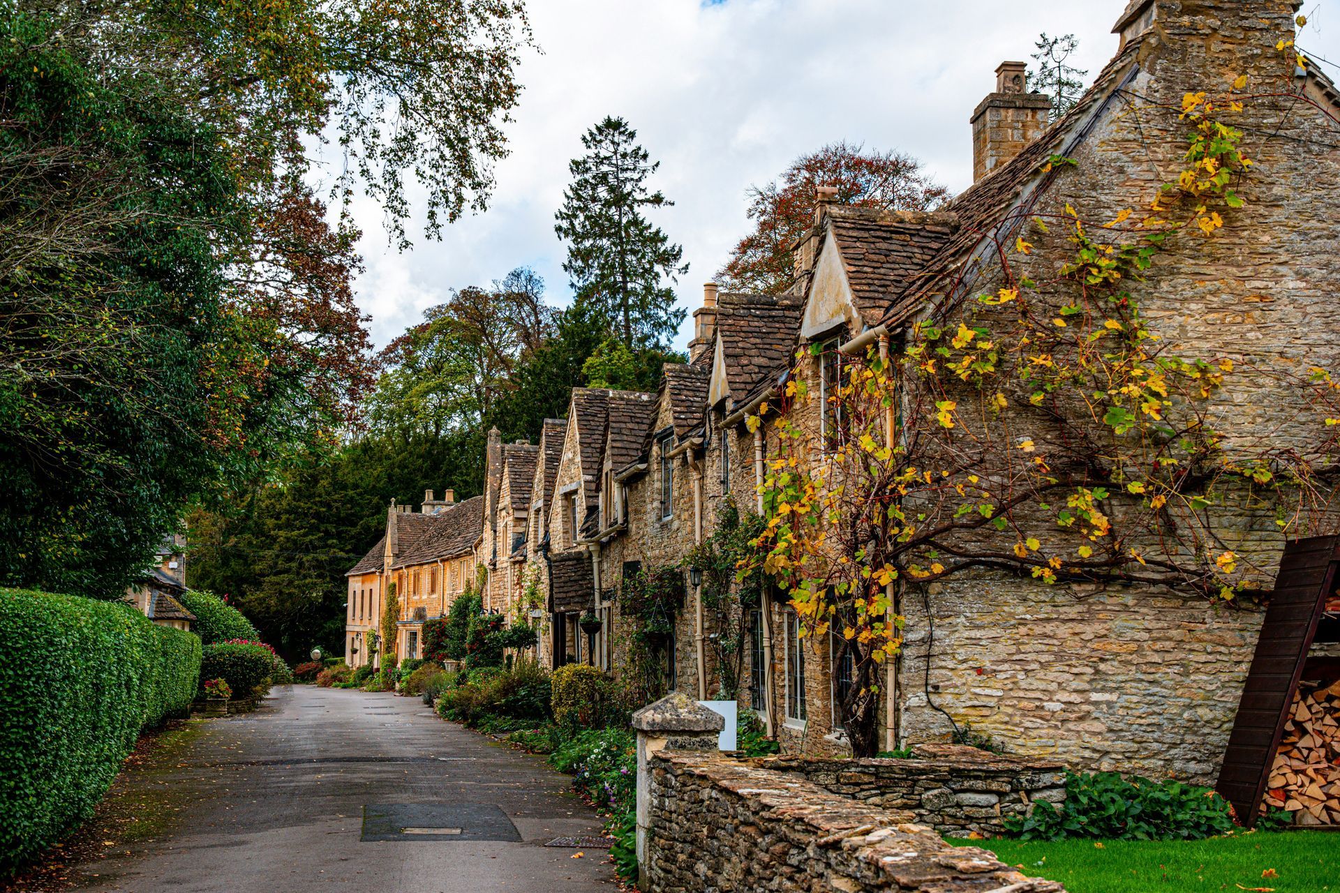 Cotswold stone cottages line a narrow road, with a stone wall and greenery in the Cotswolds, England.