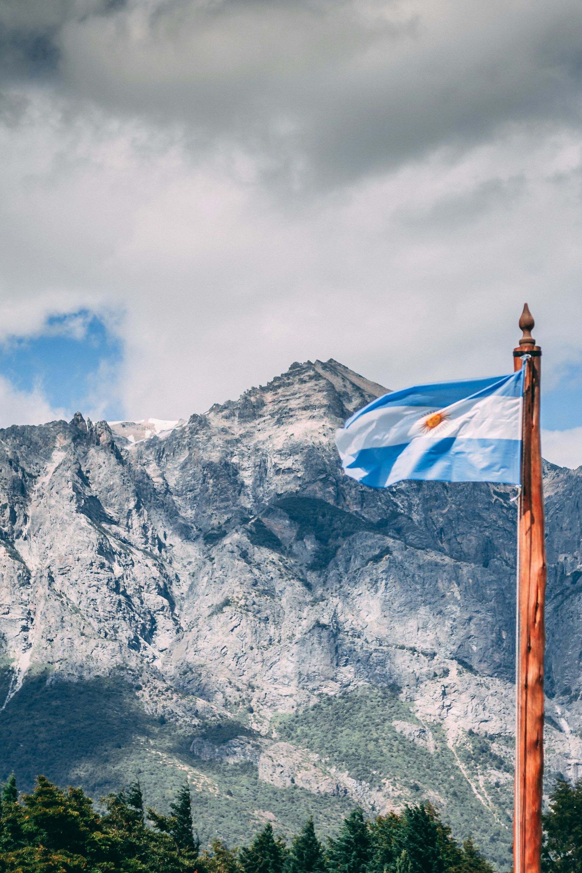 Argentine flag waves in front of a rugged, snowy mountain under a cloudy sky in San Carlos de Bariloche, Argentina.