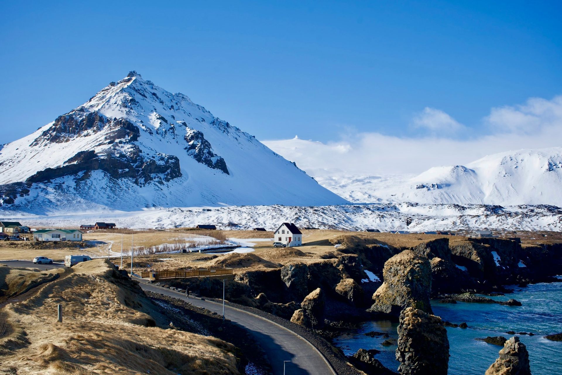 Snowy mountain overlooking a coastline with a road, small buildings, and clear blue sky in Snæfellsnes, Iceland.