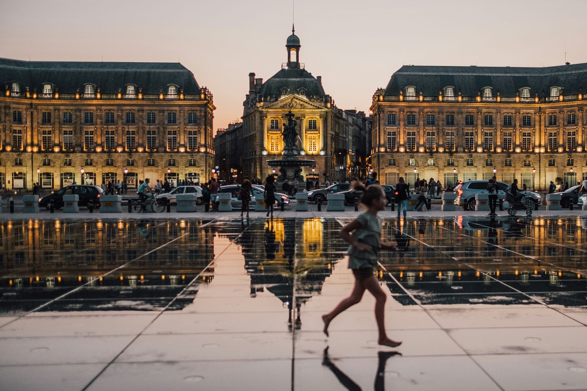 A woman is walking on a wet sidewalk in front of the Place de la Bourse in Bordeaux, France.