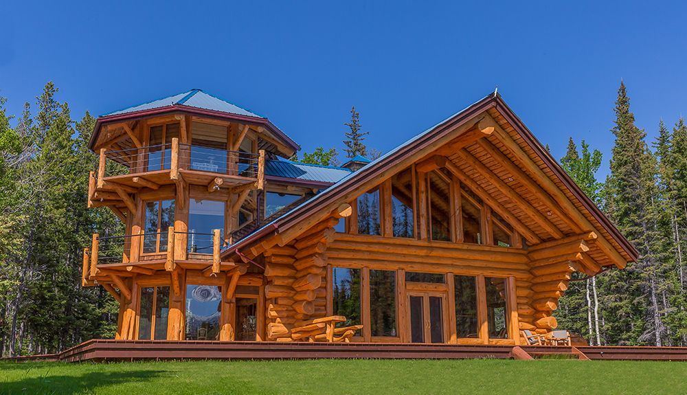 A large log cabin is sitting on top of a lush green field surrounded by trees on Chilko Lake, Canada.