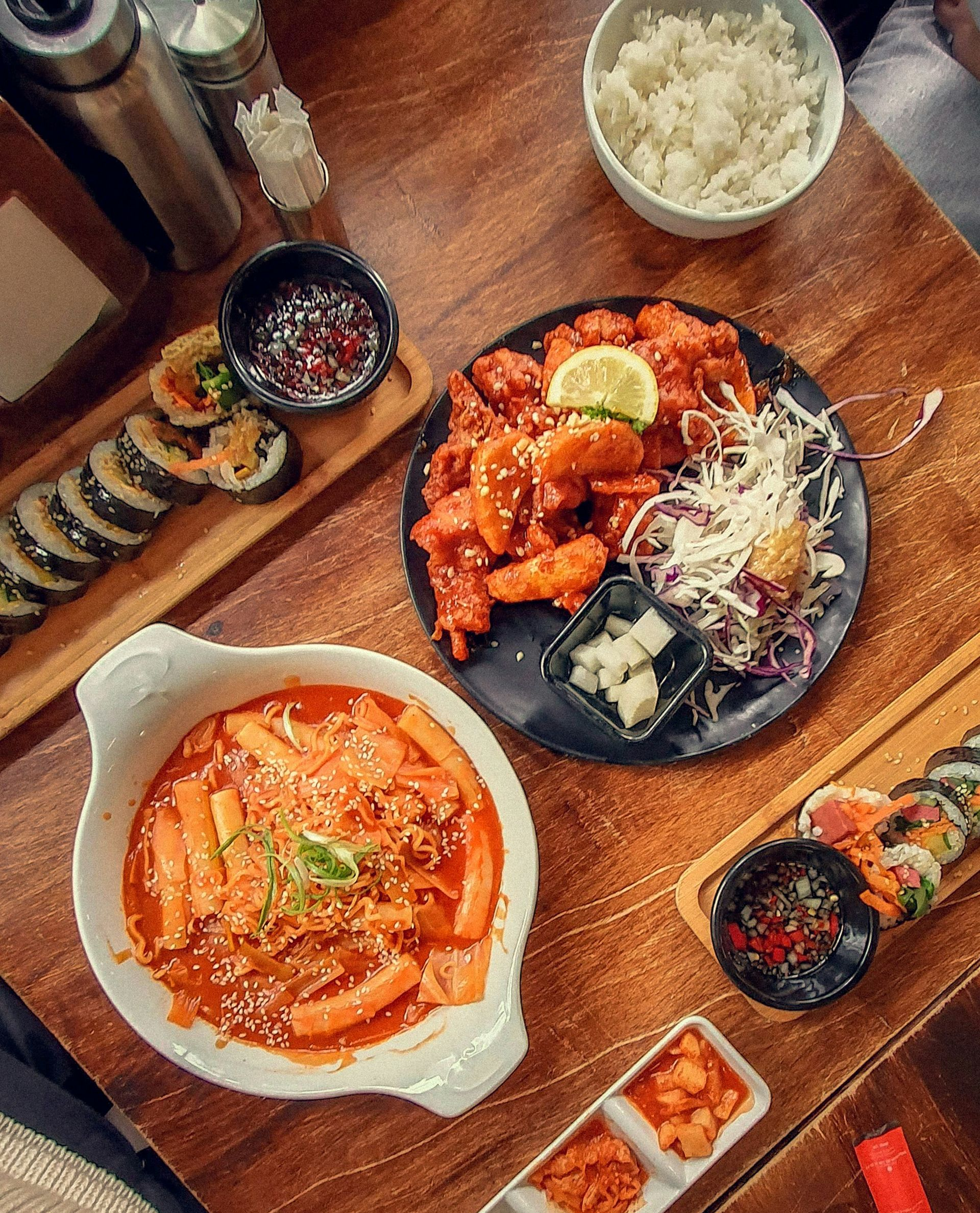 Overhead shot of Korean food spread on a wooden table: tteokbokki, fried chicken, kimbap, rice, and kimchi.