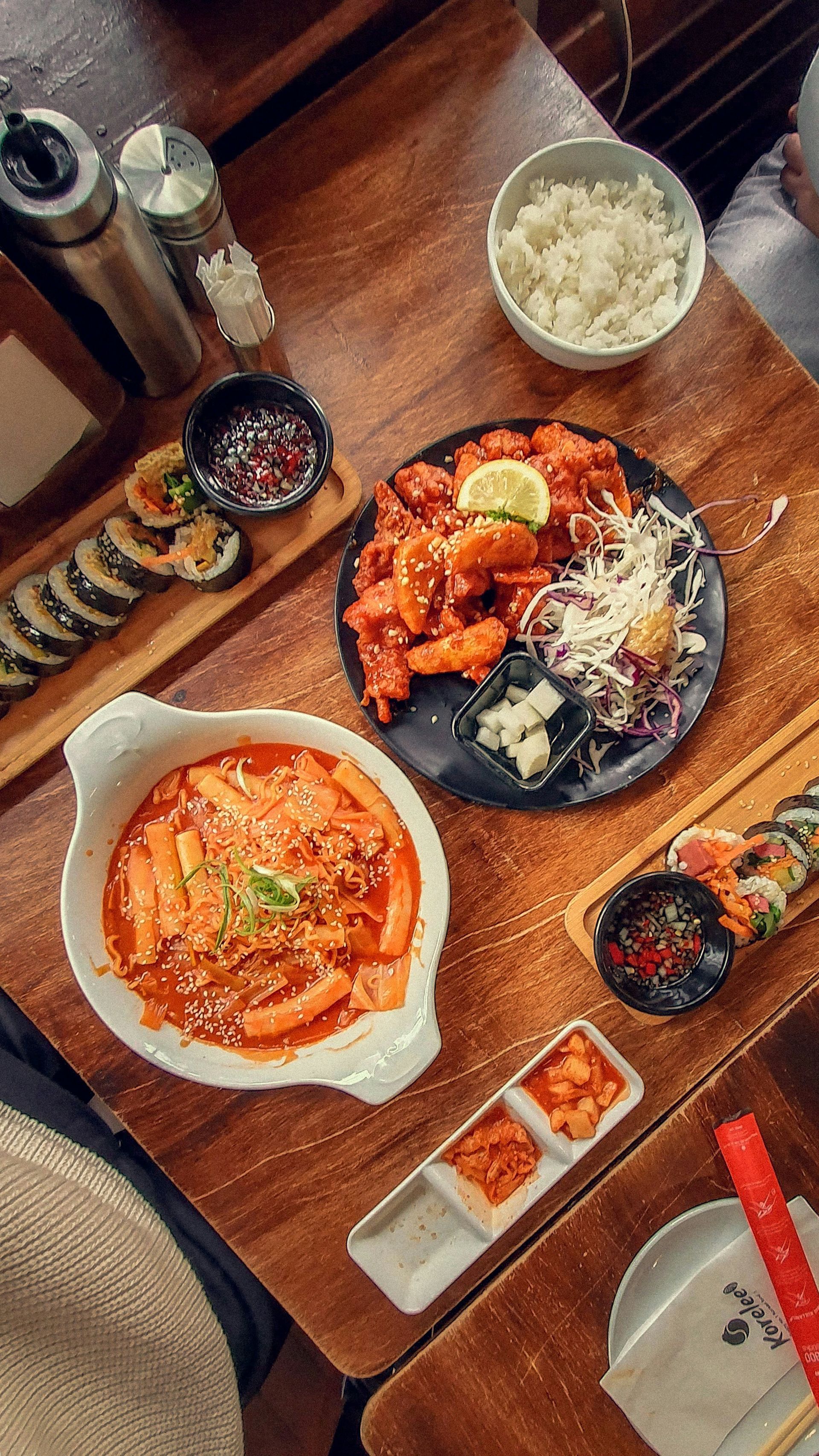Overhead shot of Korean food spread on a wooden table: tteokbokki, fried chicken, kimbap, rice, and kimchi.