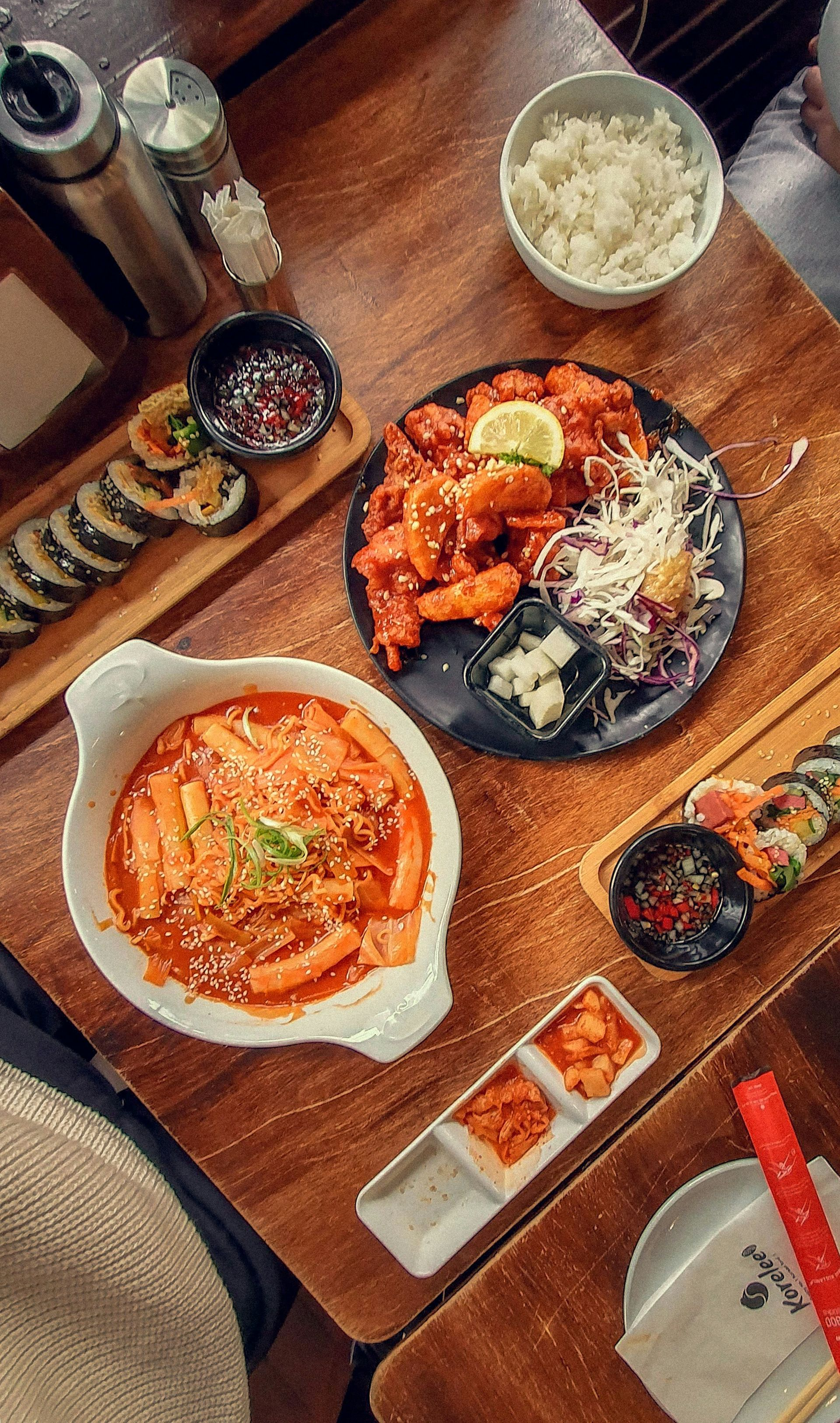Overhead shot of Korean food spread on a wooden table: tteokbokki, fried chicken, kimbap, rice, and kimchi.