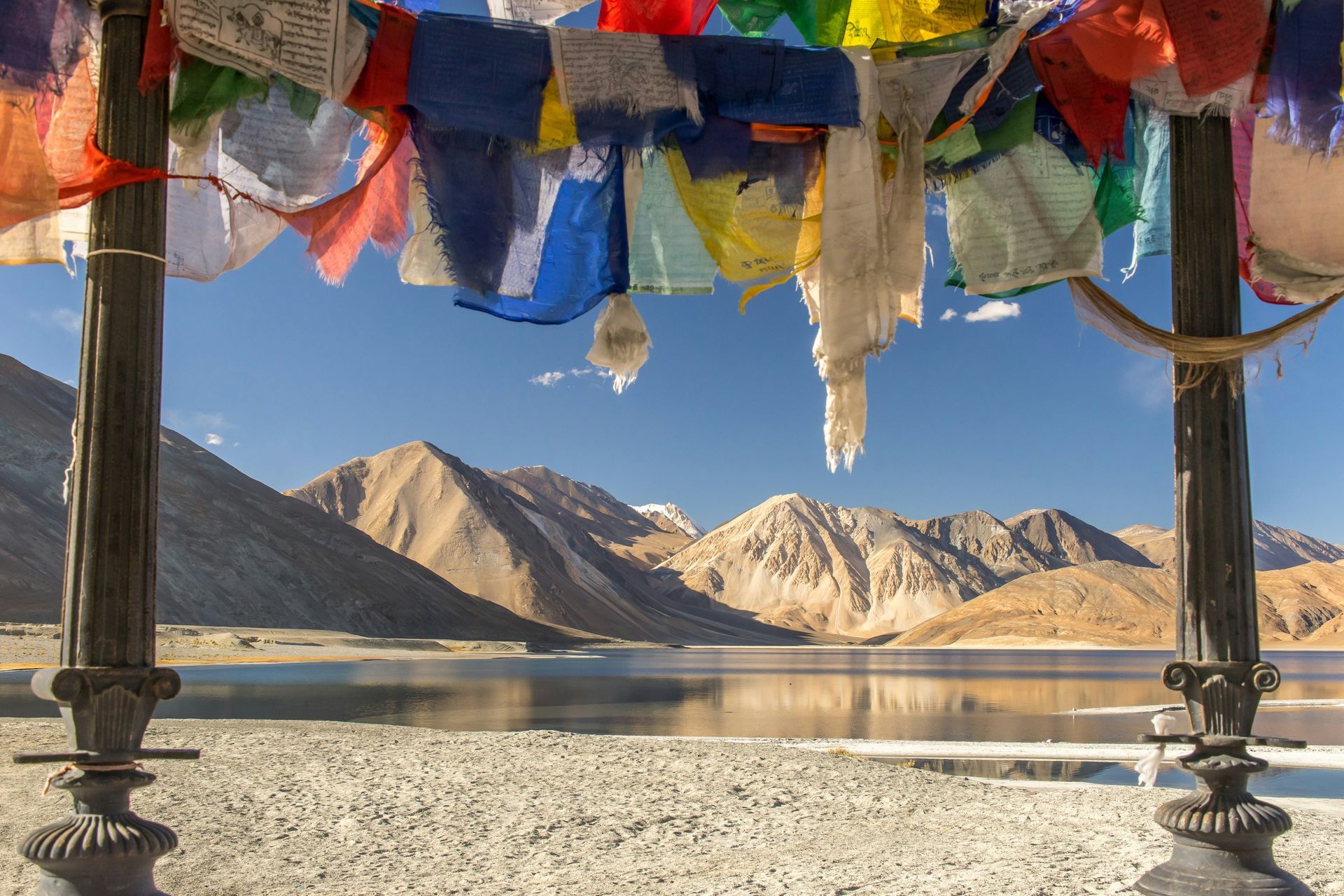 Colorful prayer flags frame a serene view of a lake and mountains in Ladakh, India.