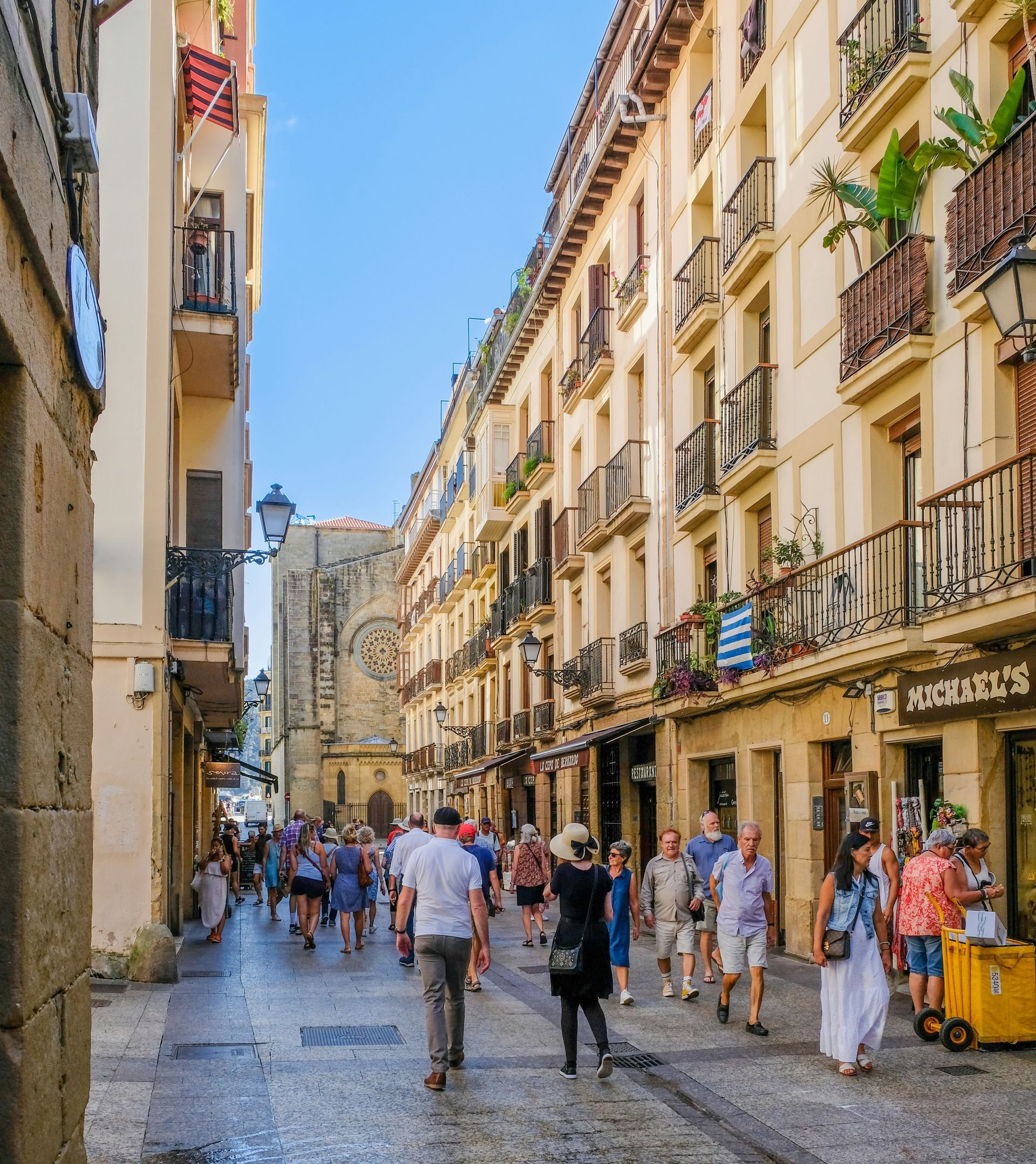 A crowded street in San Sebastián, Spain with pedestrians and buildings. Sunlight highlights the buildings.